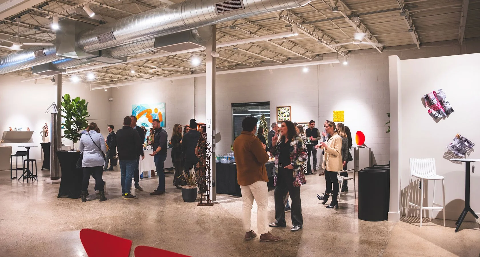 People mingling and chatting at an indoor art gallery or exhibition space with white walls, artwork on display, potted plants, and modern furniture.