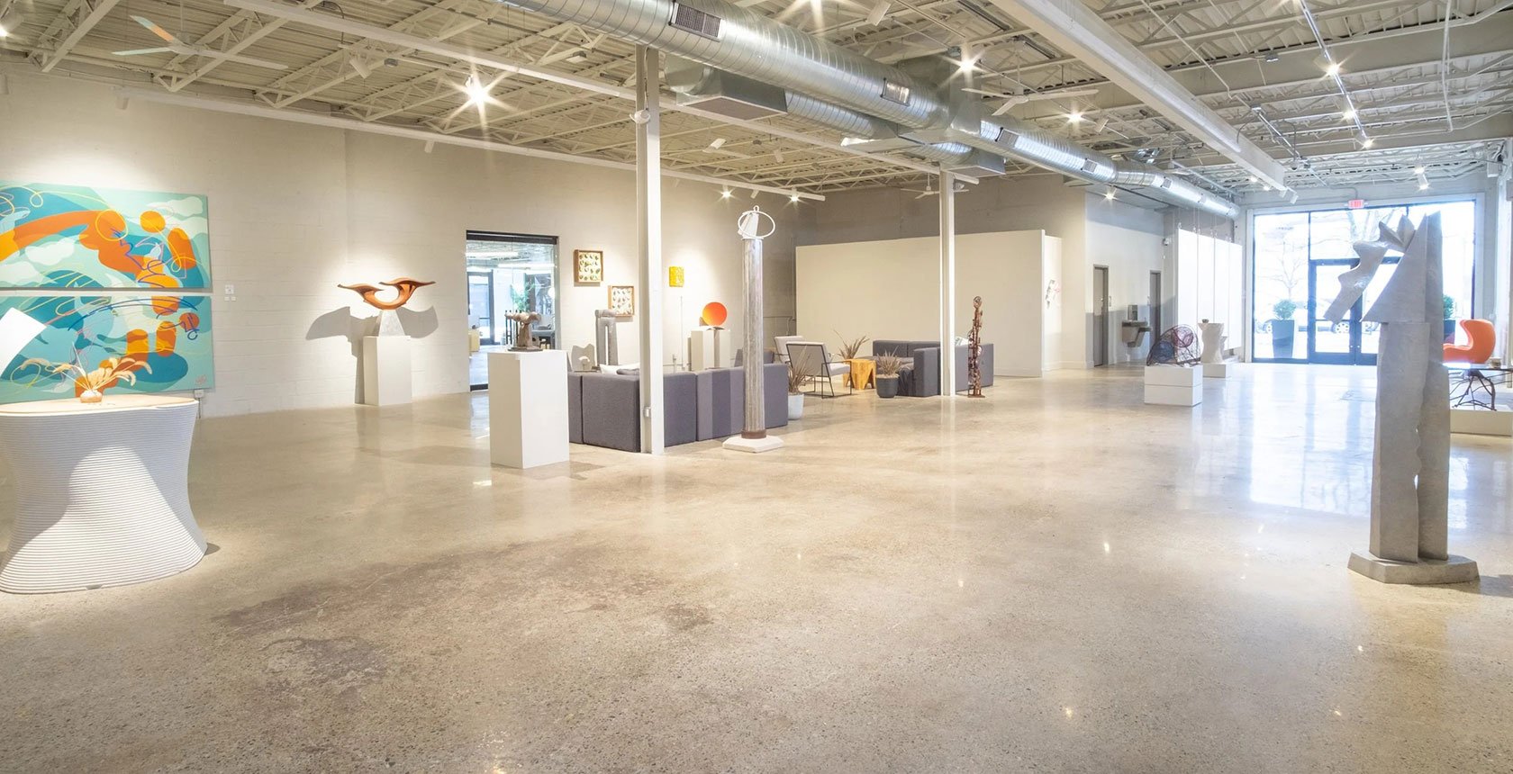 A spacious art gallery with various sculptures, paintings, and modern furniture, illuminated by ceiling lights and near large windows.