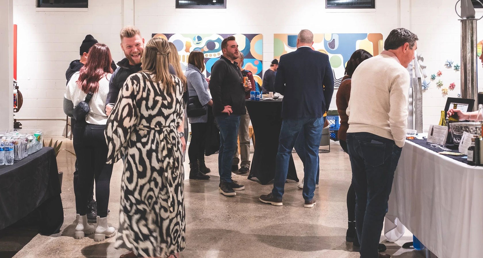 People gathered at a social event in an indoor space, with some engaging in conversation and others standing near tables with snacks and beverages, decorated with colorful artwork on the wall.