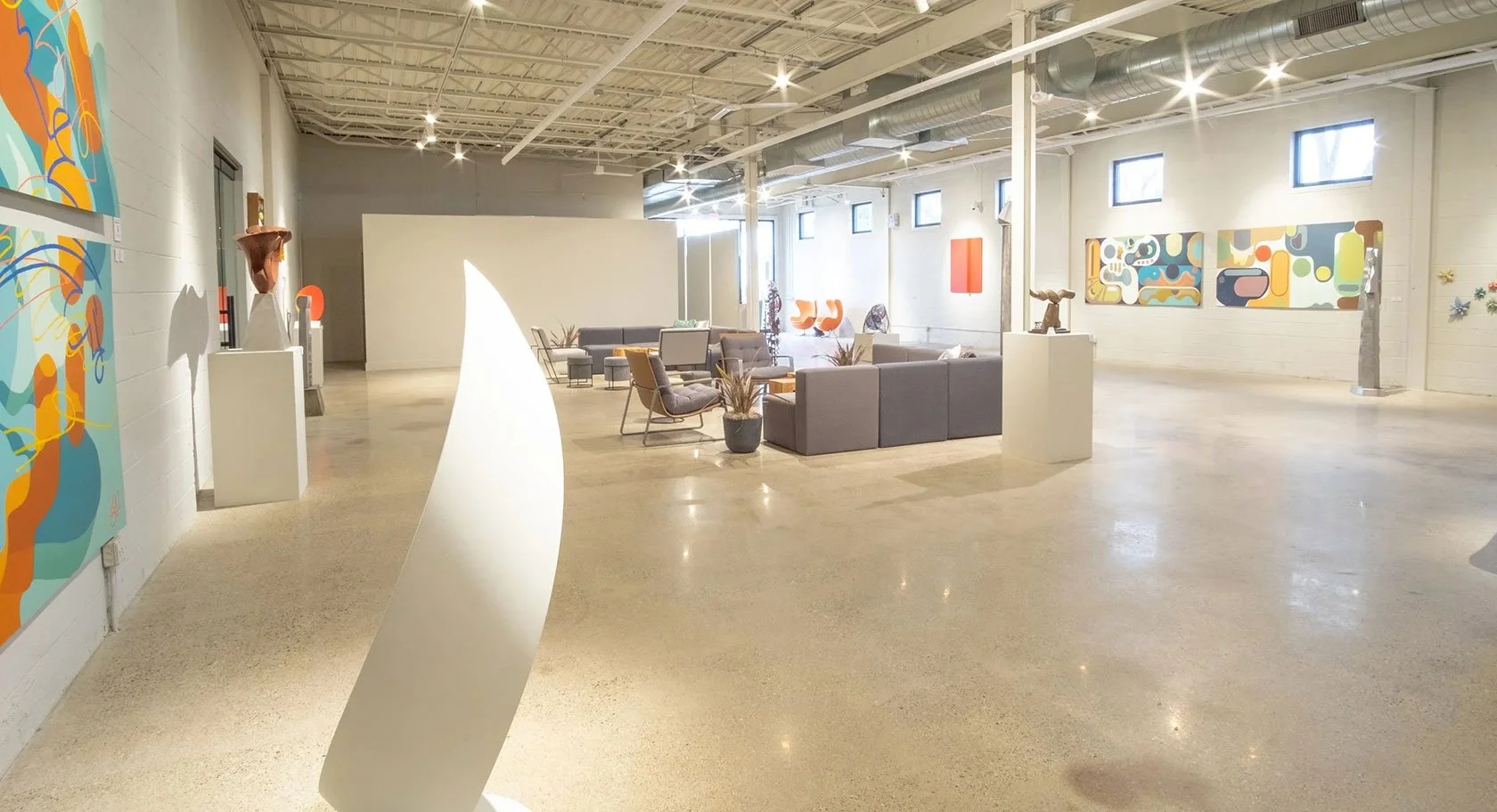 Interior of an art gallery with abstract paintings on white walls, modern furniture, sculptures, and natural lighting from small high windows.