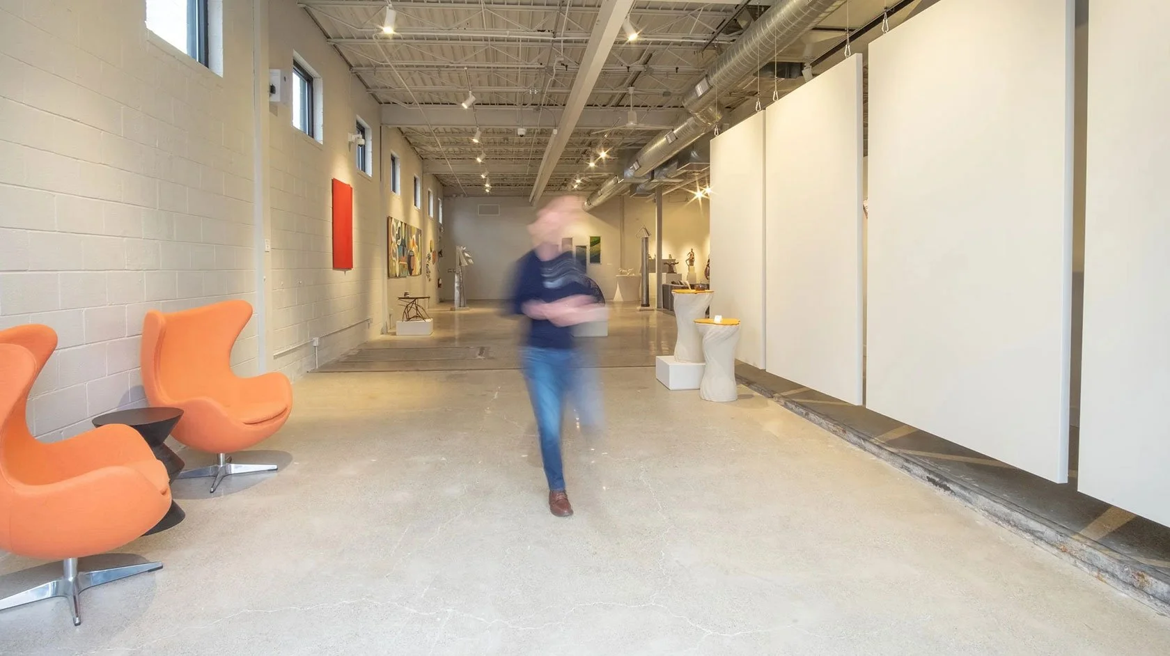 Interior of an art gallery with orange chairs on the left, white walls with paintings, and sculptures displayed in the background. A person is walking through the space, slightly blurred.