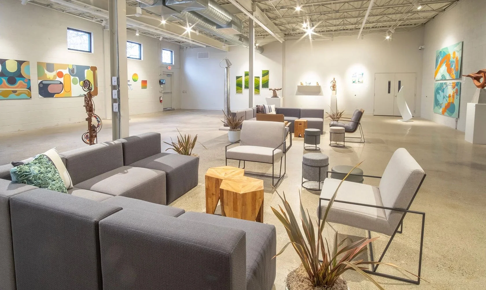 An interior view of an art gallery with colorful abstract paintings on the walls, modern furniture including sofas and chairs, potted plants, sculptures, and track lighting highlighting the artwork.