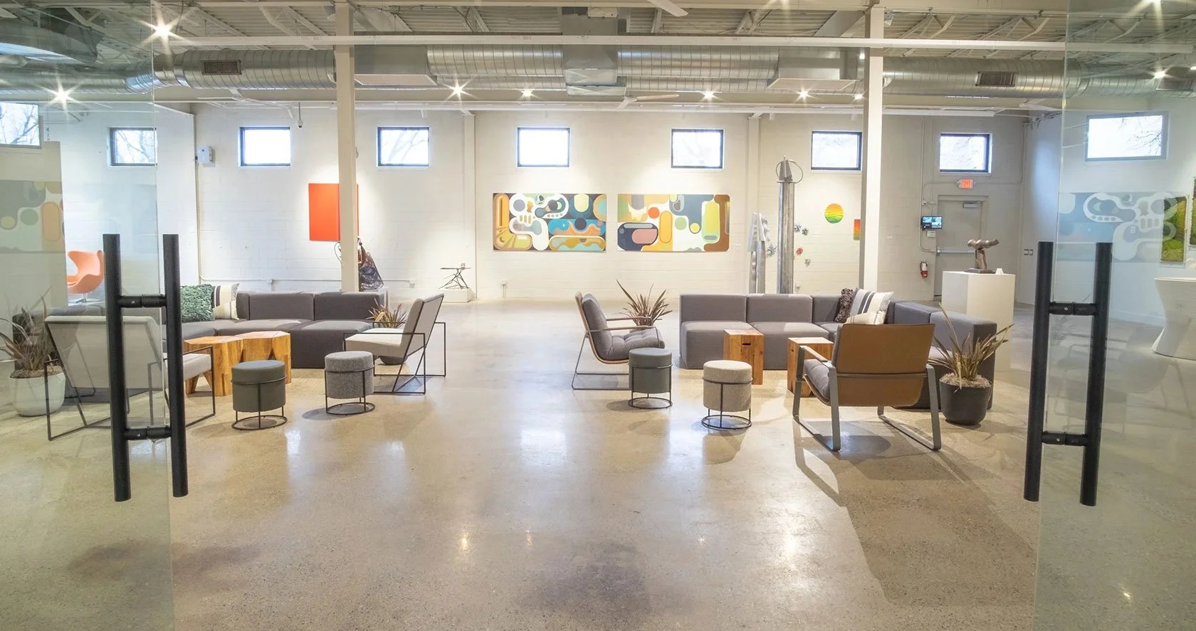 View through glass doors into a modern lounge area with sofas, chairs, small tables, and framed colorful abstract artwork on the wall.