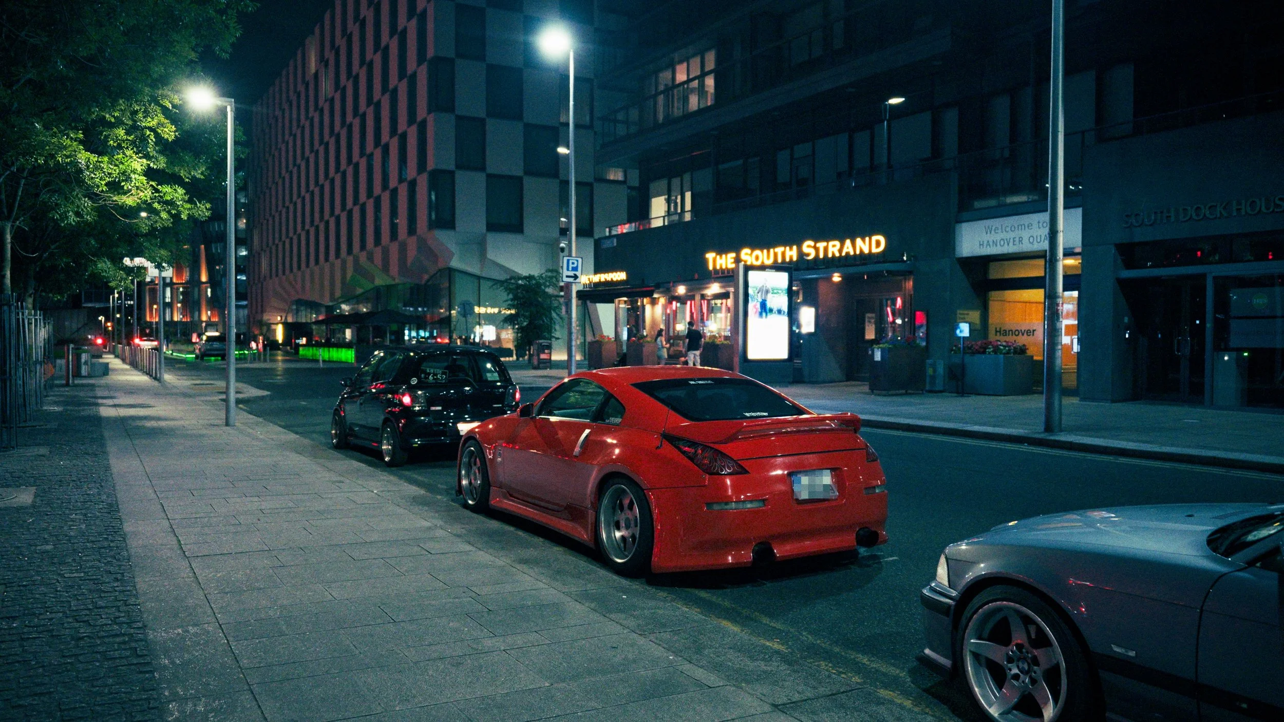Nighttime street scene showing cars parked along the sidewalk, a modern building with illuminated signs and streetlights, with a sign that says "The South Strand."