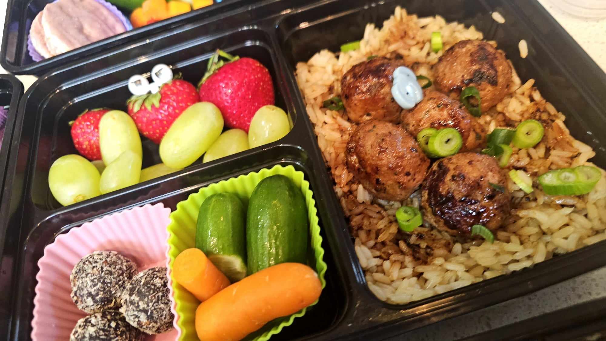 A bento style food prep container with three compartments. The compartments contain brown rice topped with meatballs, strawberries and grapes, coconut date balls in a pink silicone cup and and cucumber and carrots in a green silicone cup.