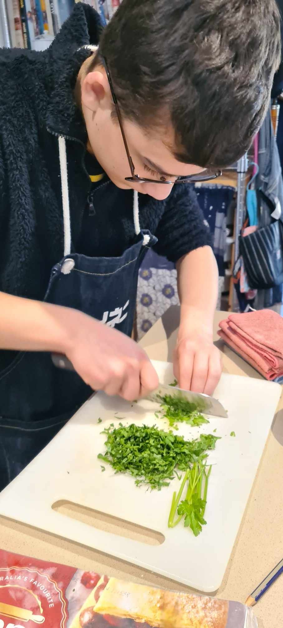Teenage boy wearing a navy jumper and apron. He is finely chopping fresh coriander on a white chopping board.