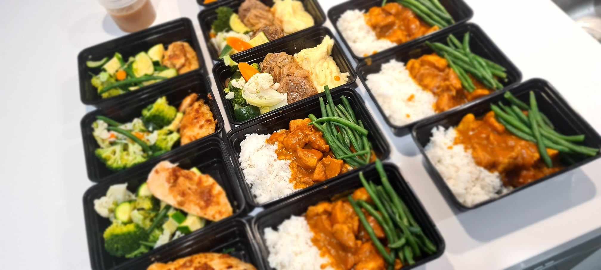 A variety of 12 single serve pre-prepared meals presented in an aesthetically pleasing way. The meals are packaged in individual black food containers.