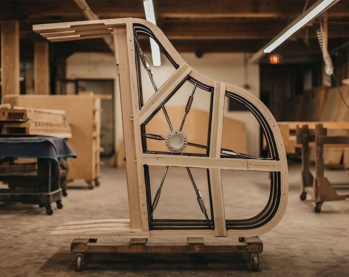 Side view of a grand piano frame being constructed inside a workshop, showing wooden beams and metal components.
