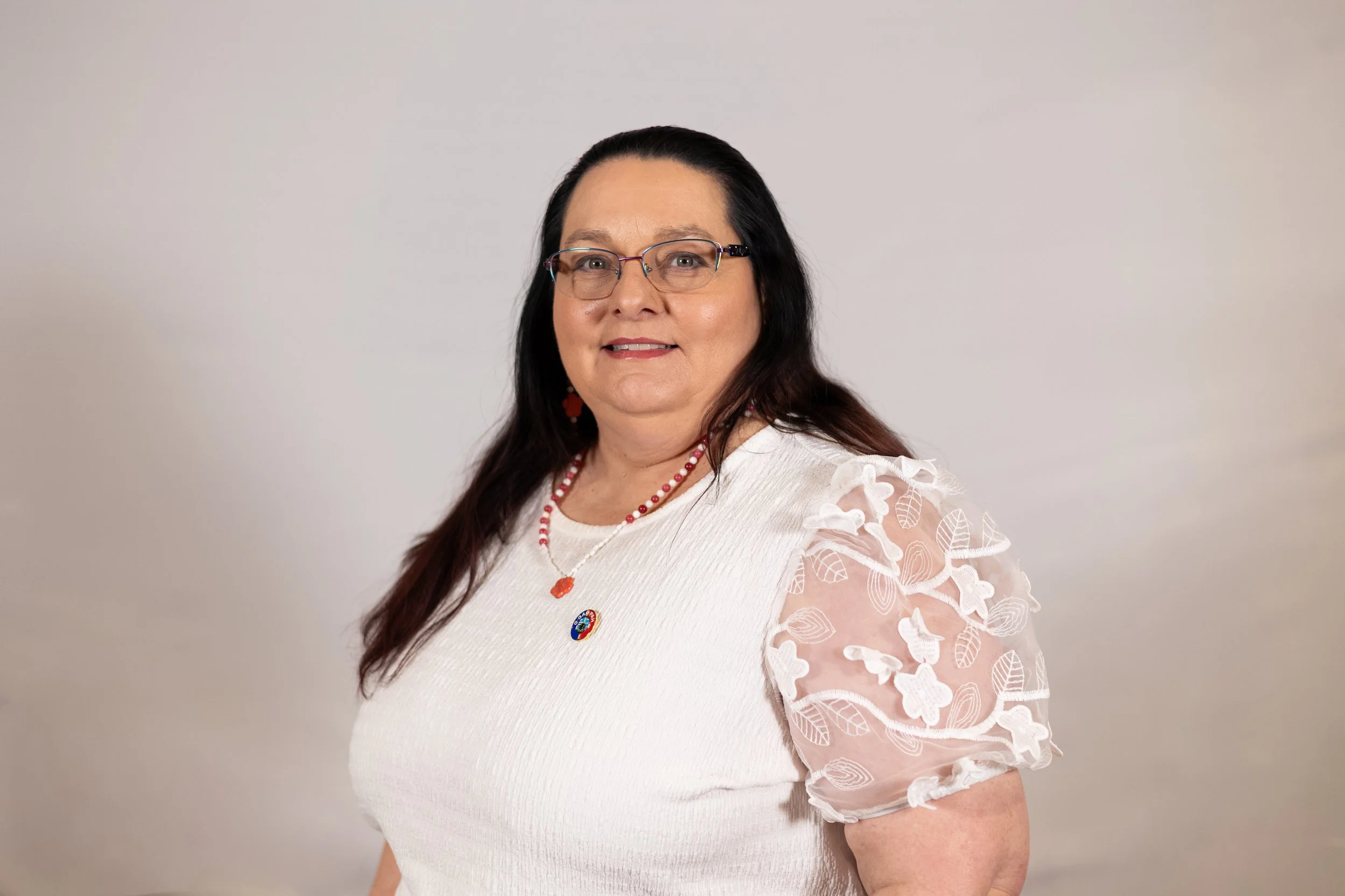 Linda Valliere wearing a white top with embroidered leaf patterns on the sleeves, layered jewelry including a red beaded necklace and a pin on her chest.