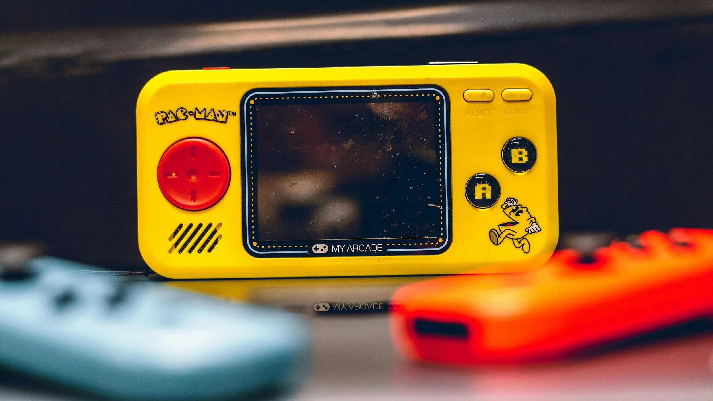 Yellow handheld electronic game with Pac-Man graphics, featuring a red directional pad, black and yellow buttons, and a small screen in the center, placed on a surface with blurred objects in the foreground.