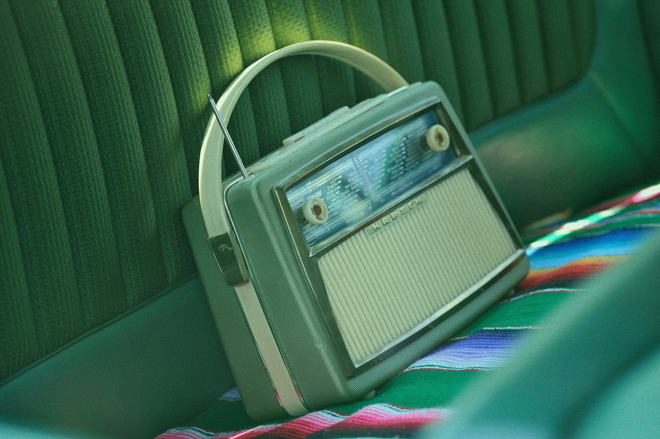 A vintage portable radio sitting on a green seat with a colorful striped fabric underneath, against a green textured wall.