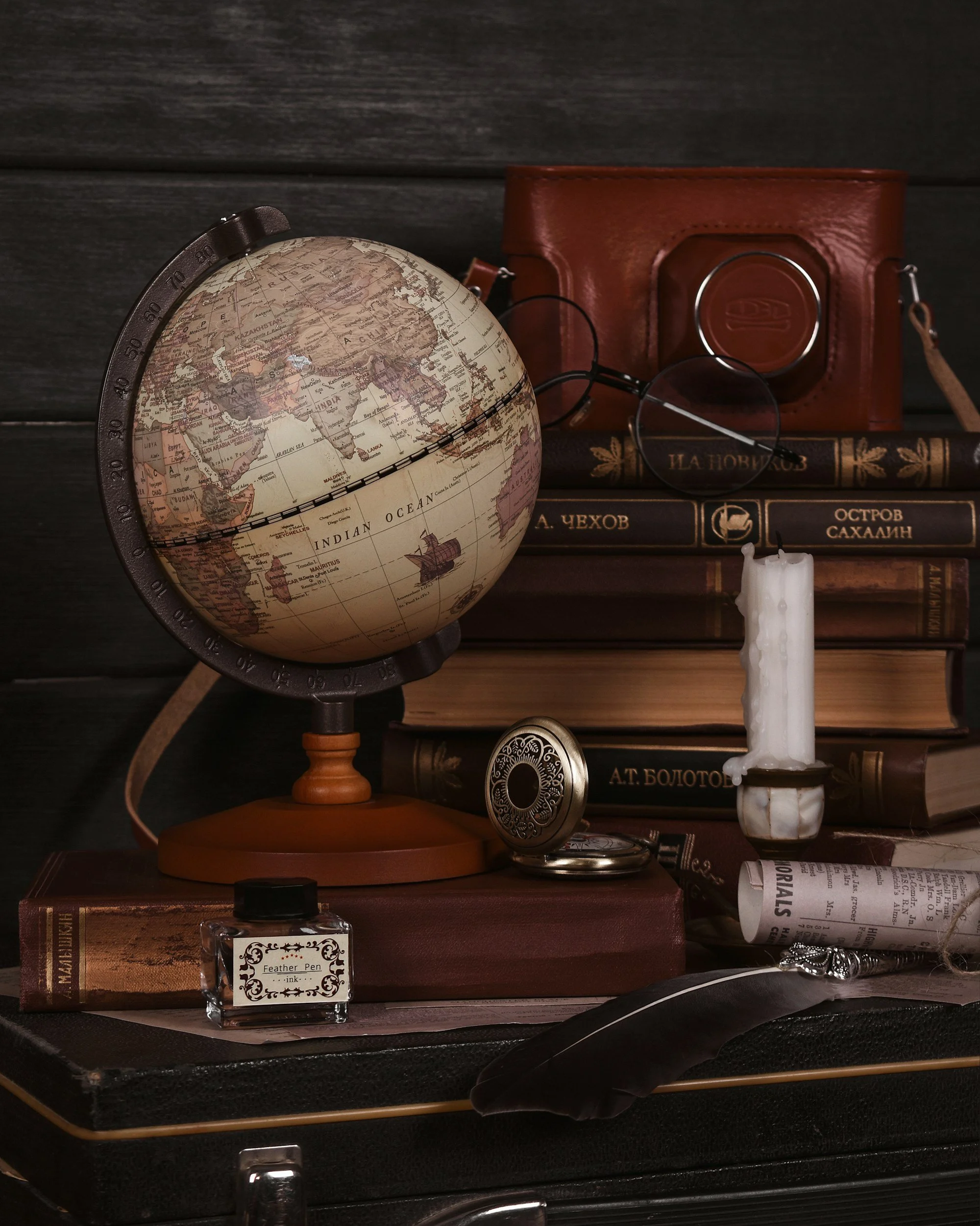 A vintage globe, a leather camera case, a magnifying glass, a stack of books with Russian titles, a white candle, a rolled newspaper, a small ink bottle labeled 'Feather Pen ink', a pocket watch, and a black feather quill, all arranged on a dark textured surface with a dark wooden background.