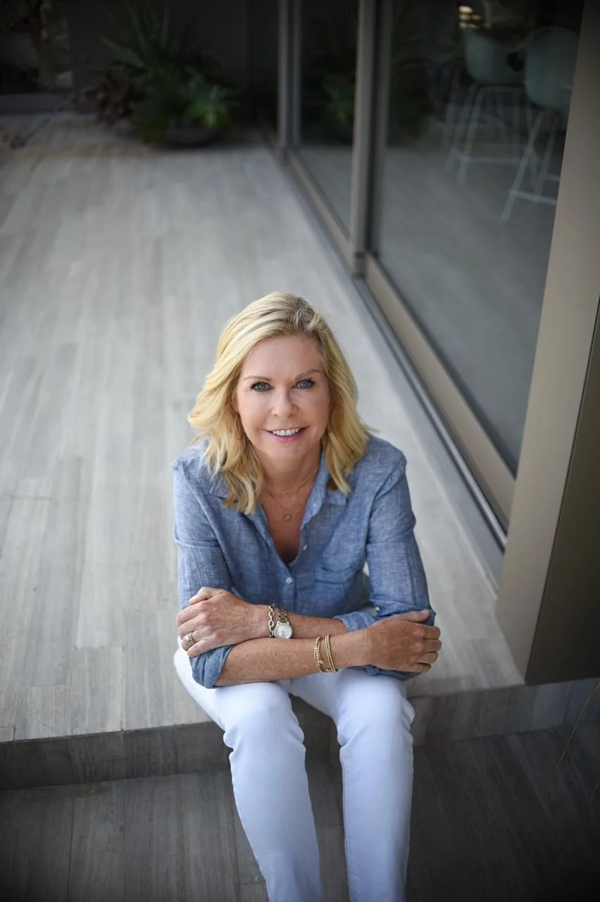 Laurie James, wearing a linen shirt and white pants, smiling and sitting on a step inside a modern home, with a sliding glass door and plants in the background.