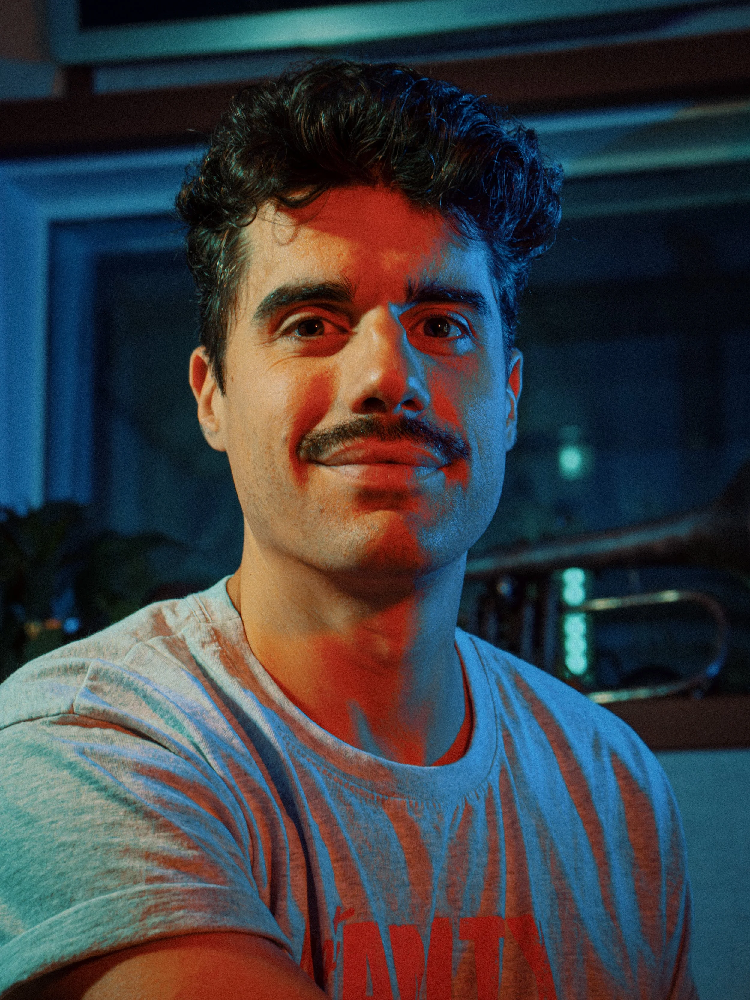 Jeune homme souriant avec un motif moustache, portant un t-shirt gris à rayures, dans un environnement avec des lumières colorées.