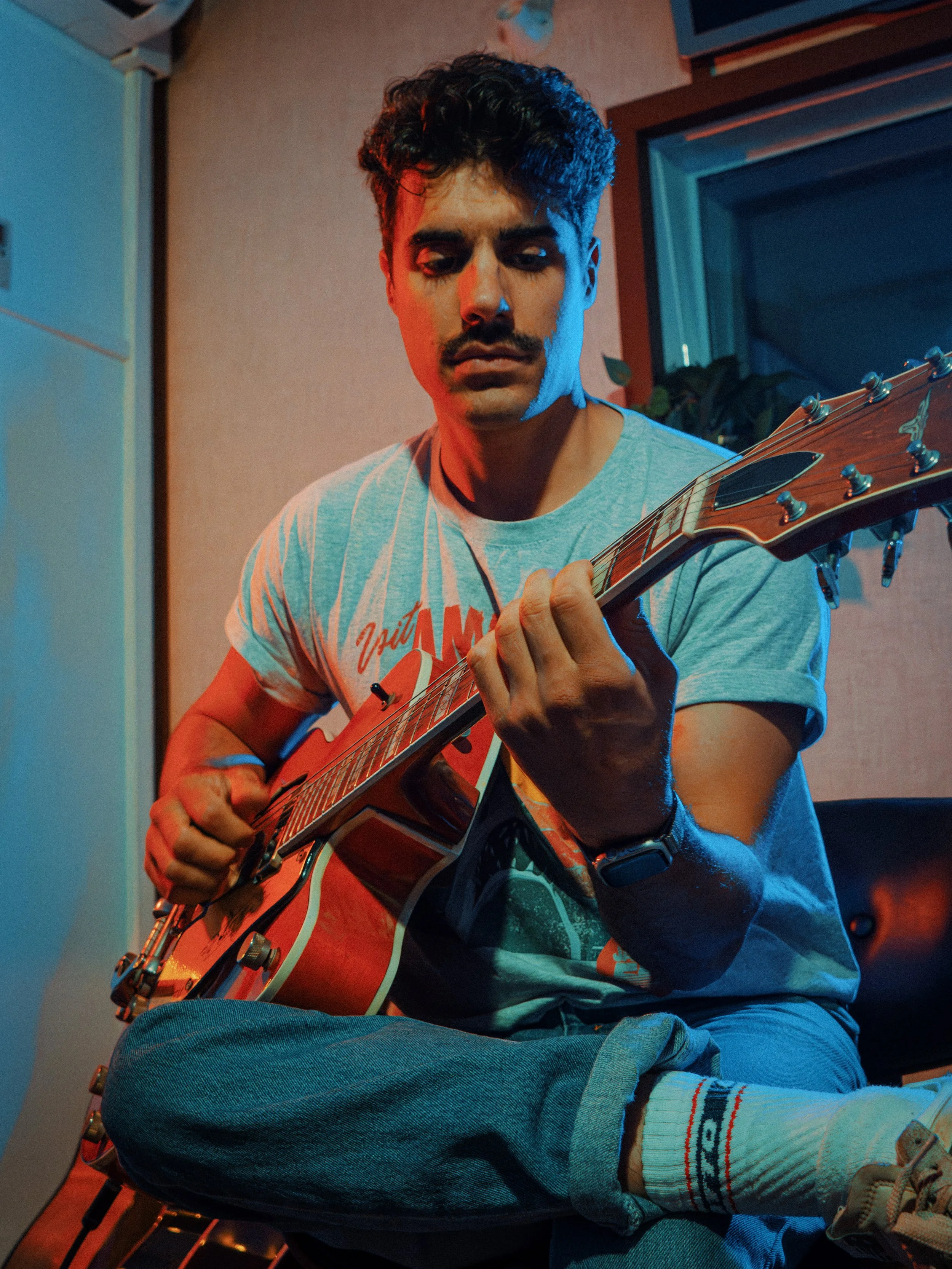 Un jeune homme assis en tailleur joue de la guitare électrique dans une pièce, éclairé par une lumière bleue et rouge.