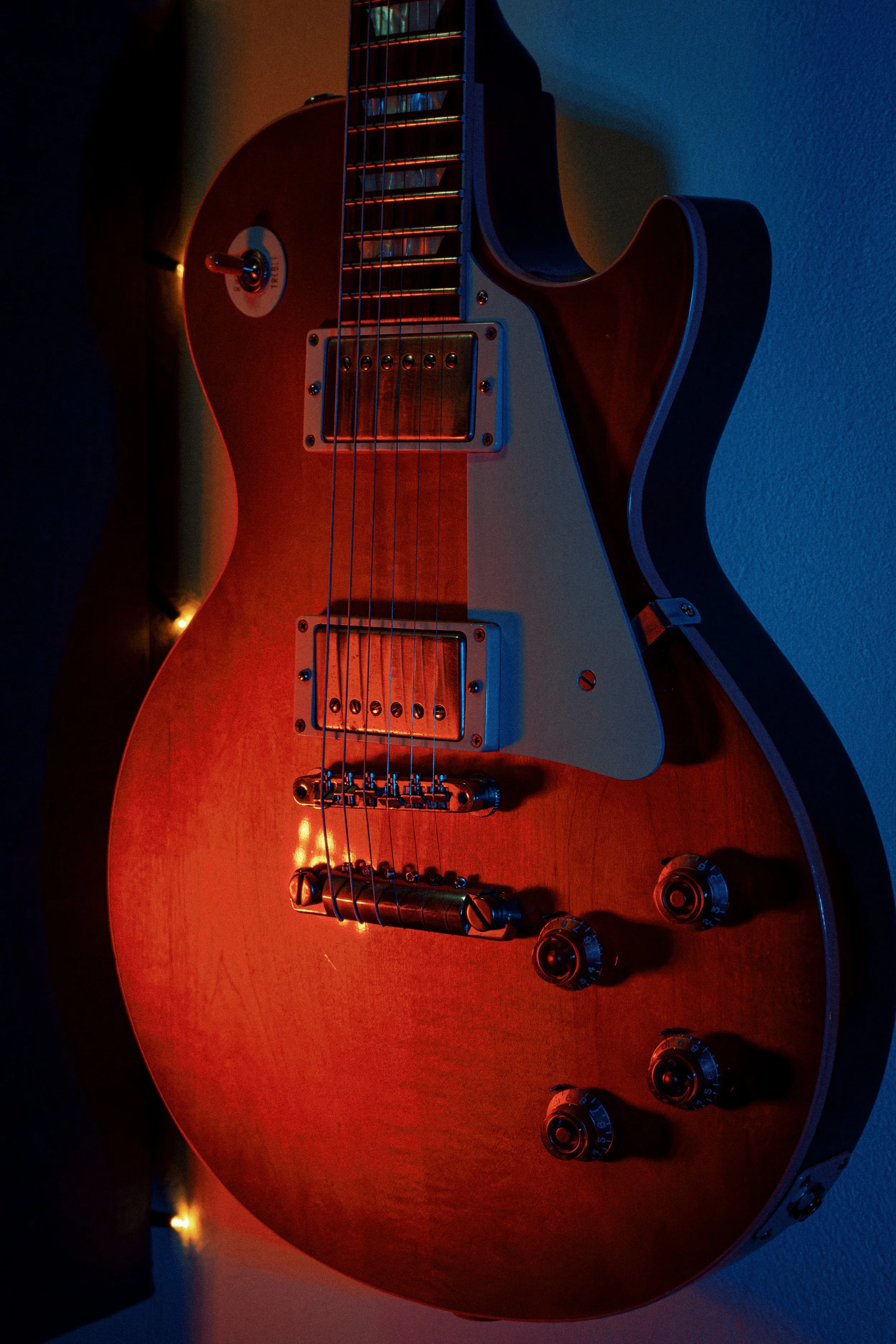 Guitare électrique en bois avec plusieurs boutons et réglages, éclairée par une lumière rouge. En arrière-plan, une autre guitare sombre sur un mur.