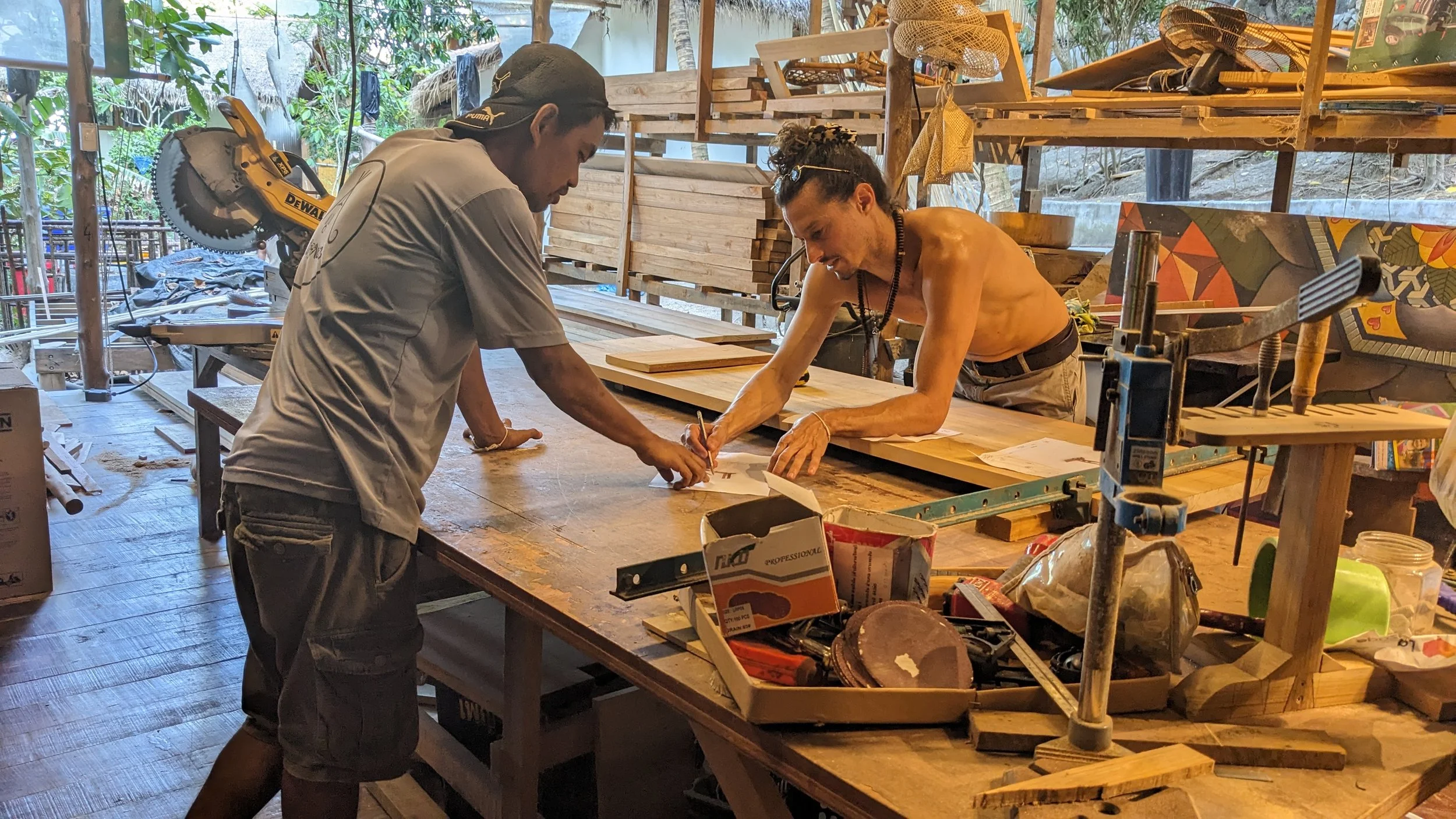 Two men are working together in a woodworking shop, examining a piece of wood on a workbench. One man is shirtless while the other is wearing a gray shirt and a dark cap. The workshop is filled with various tools, wood pieces, and supplies, and natural light is coming in through the windows and open walls.