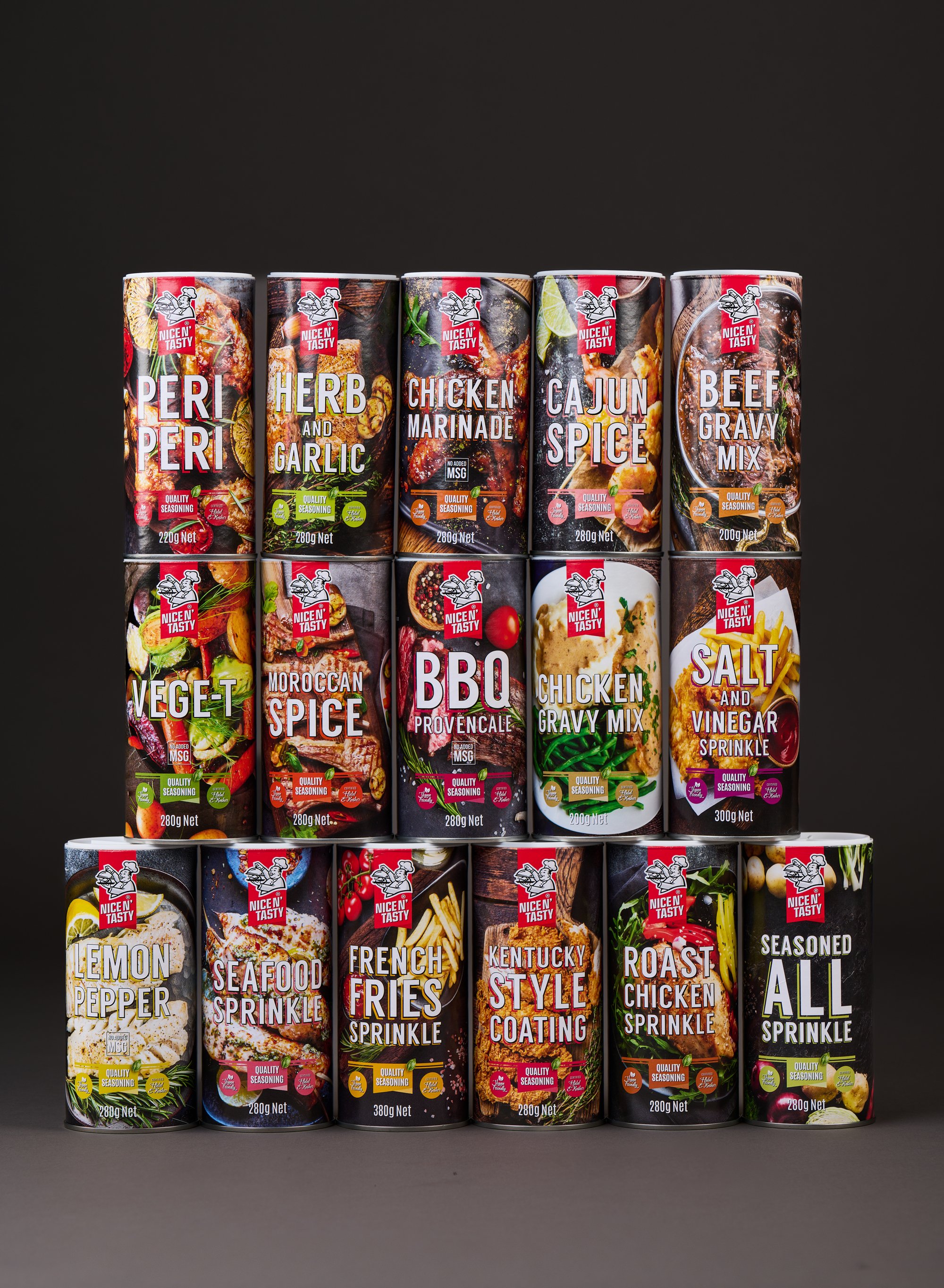 Stacked cans of various seasoning and flavoring products on a black background, including peri peri, herb and garlic, chicken marinade, cajun spice, beef gravy mix, vegetable seasoning, Moroccan spice, BBQ provencale, chicken gravy mix, salt and vinegar sprinkle, lemon pepper, seafood sprinkle, French fries sprinkle, Kentucky style coating, roast chicken sprinkle, and seasoned all sprinkle.