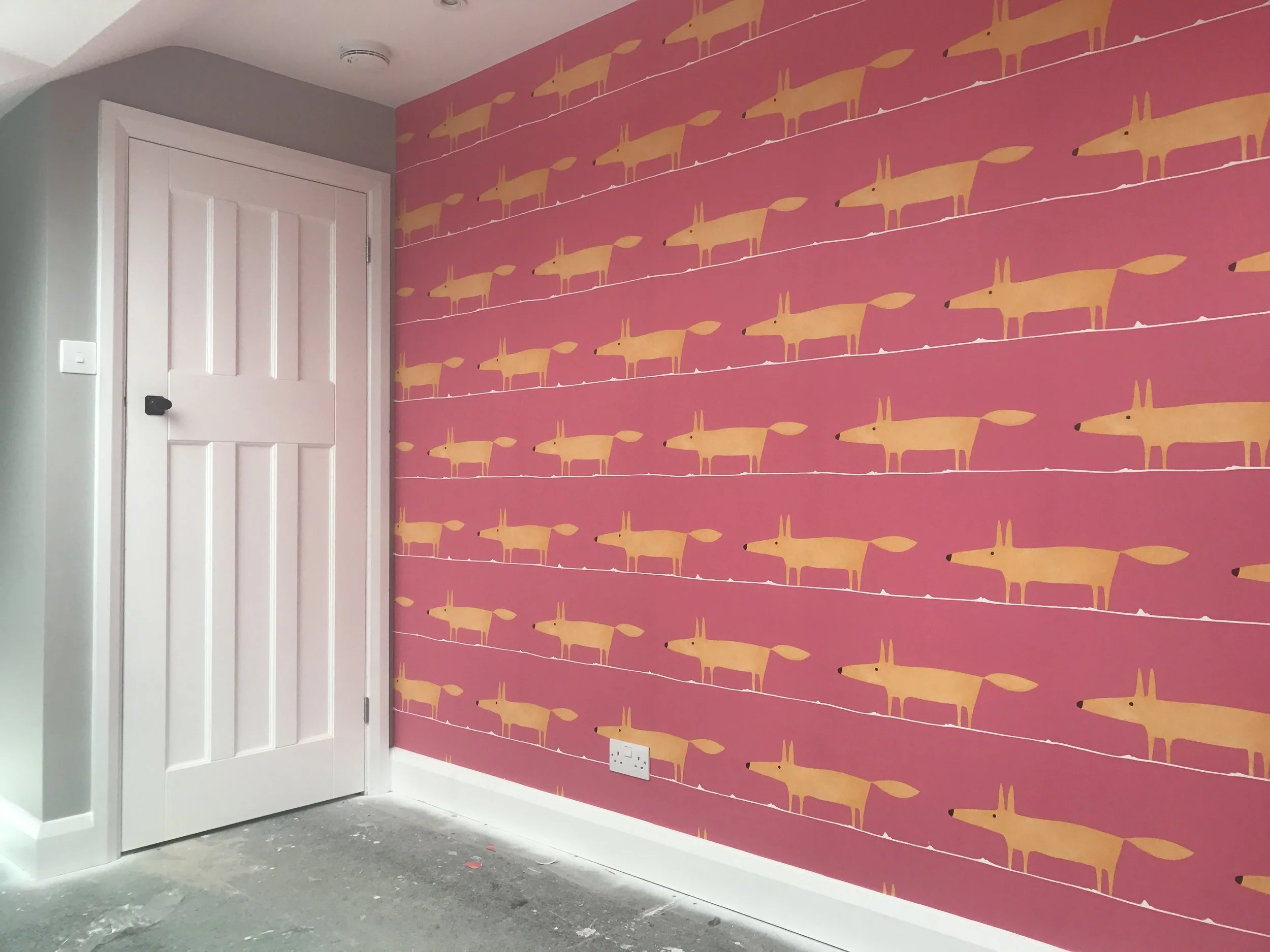 A room corner with pink wallpaper featuring a repeating pattern of yellow foxes, white horizontal lines, a white door on the left, a gray wall, and a white electrical outlet near the floor.
