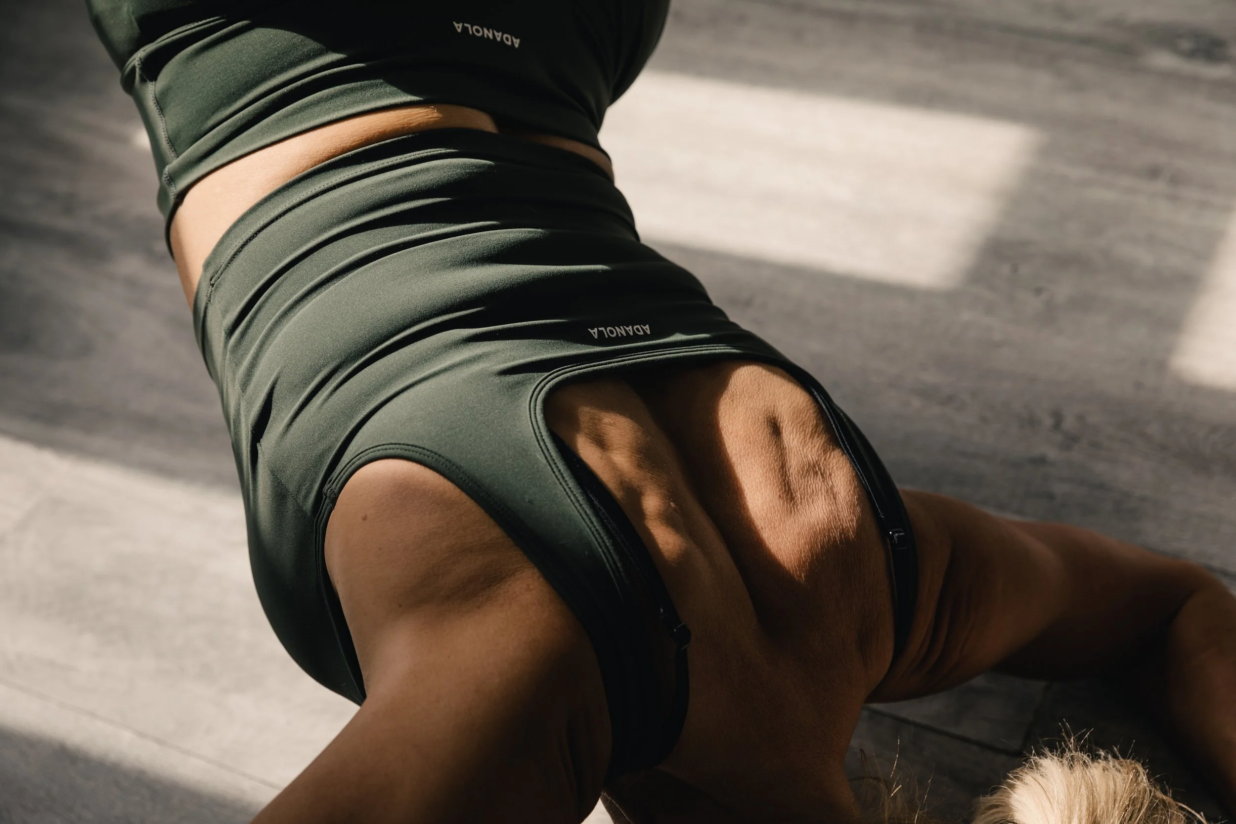 Close-up of a woman in black athletic wear doing a bridge pose on a wooden floor, natural light casting shadows.