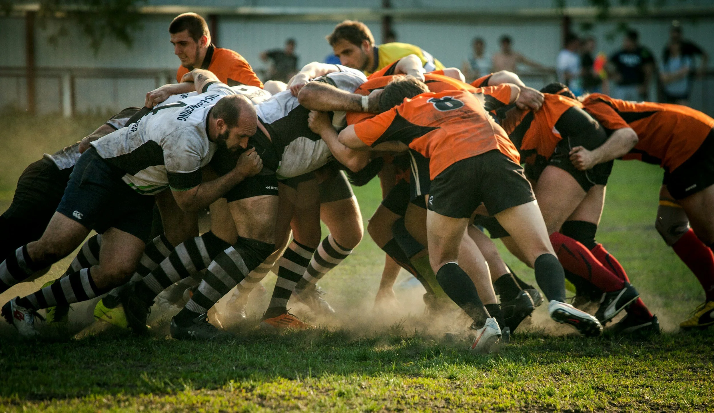 A rugby match with players engaged in a scrum on a grassy field, with spectators in the background.