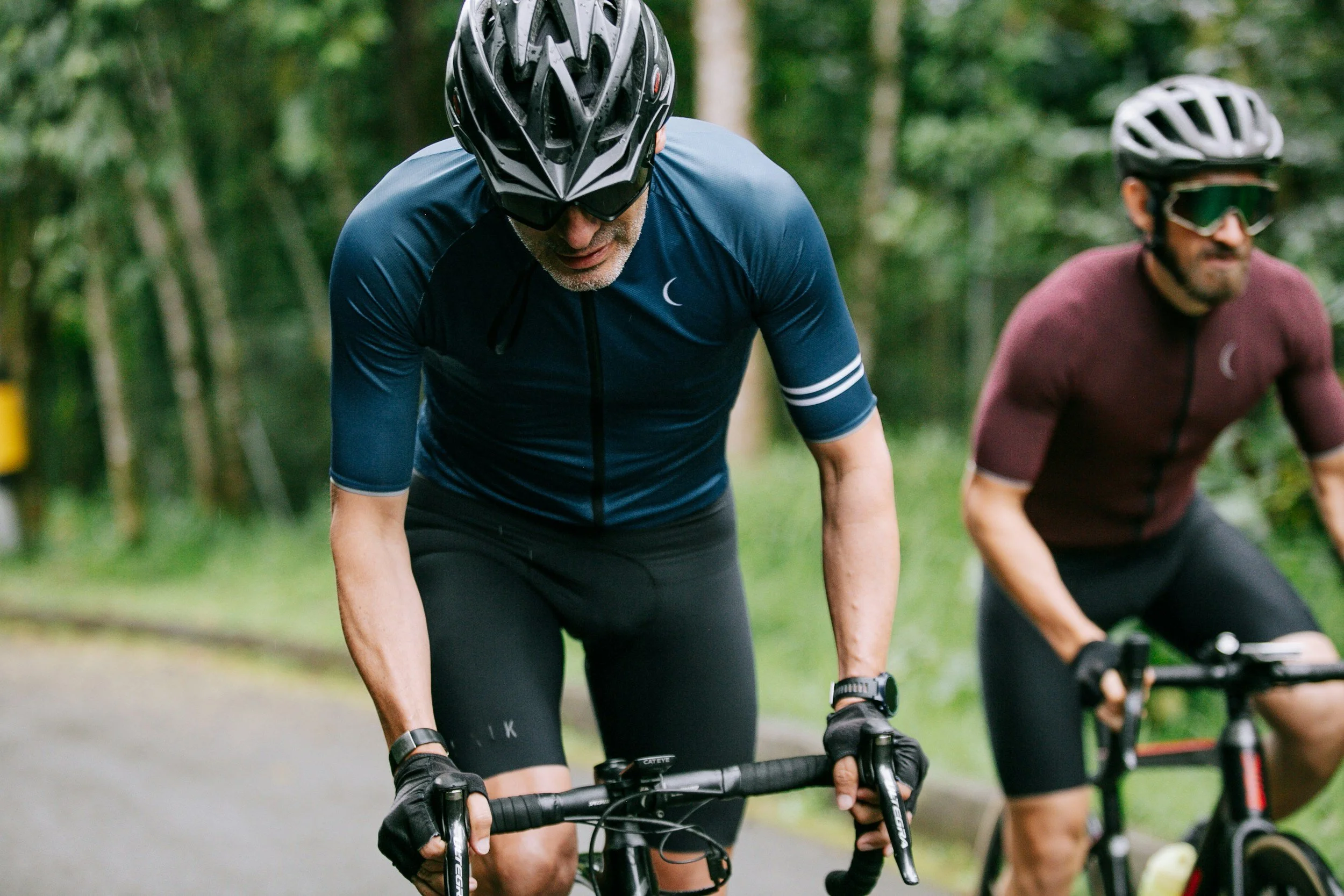 Two male cyclists wearing helmets and sportswear riding on a forest trail.