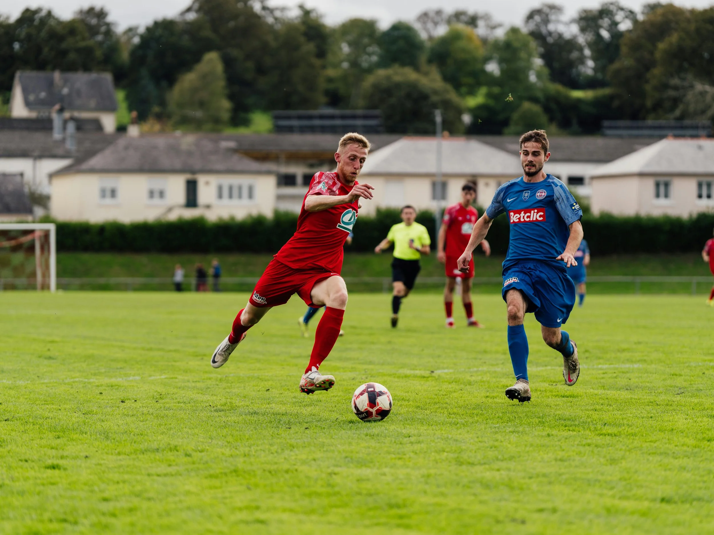football martch, 2 football players on a green field kicking a football, one in red football kit and the other in blue