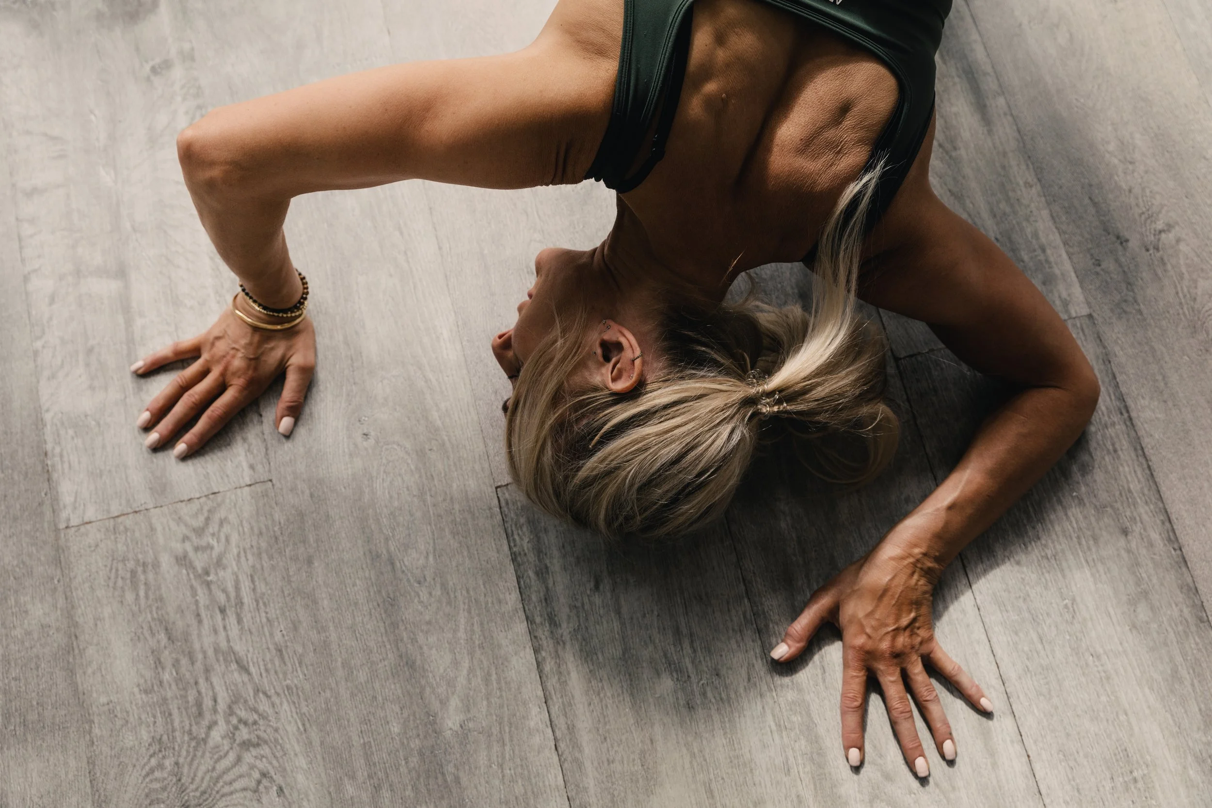 A woman practicing yoga on a wooden floor, positioned on her side with one arm extended and the other supporting her body, wearing a black sports bra and her hair in a ponytail.