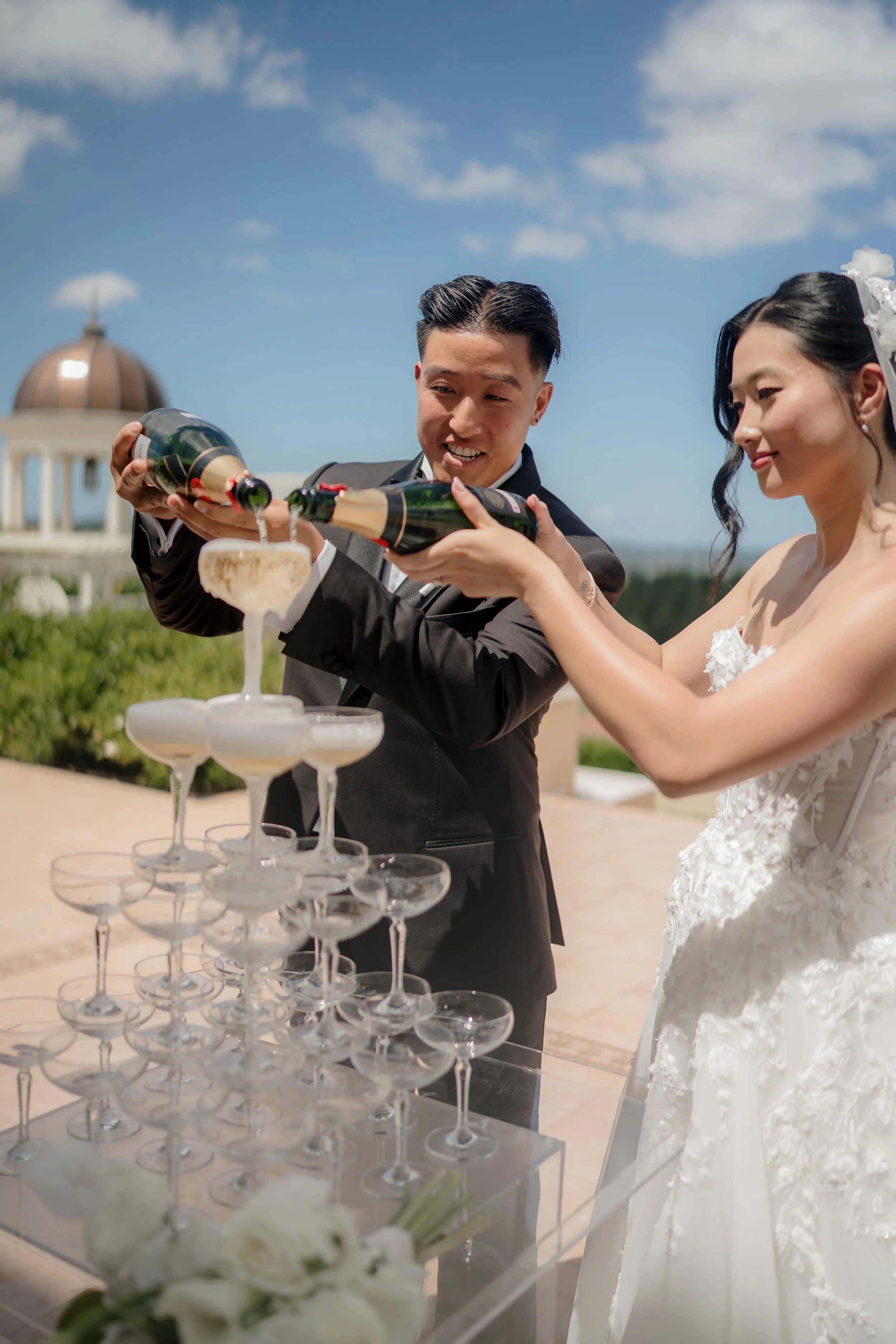A bride and groom in formal attire pouring champagne into a pyramid of glasses at a wedding celebration.