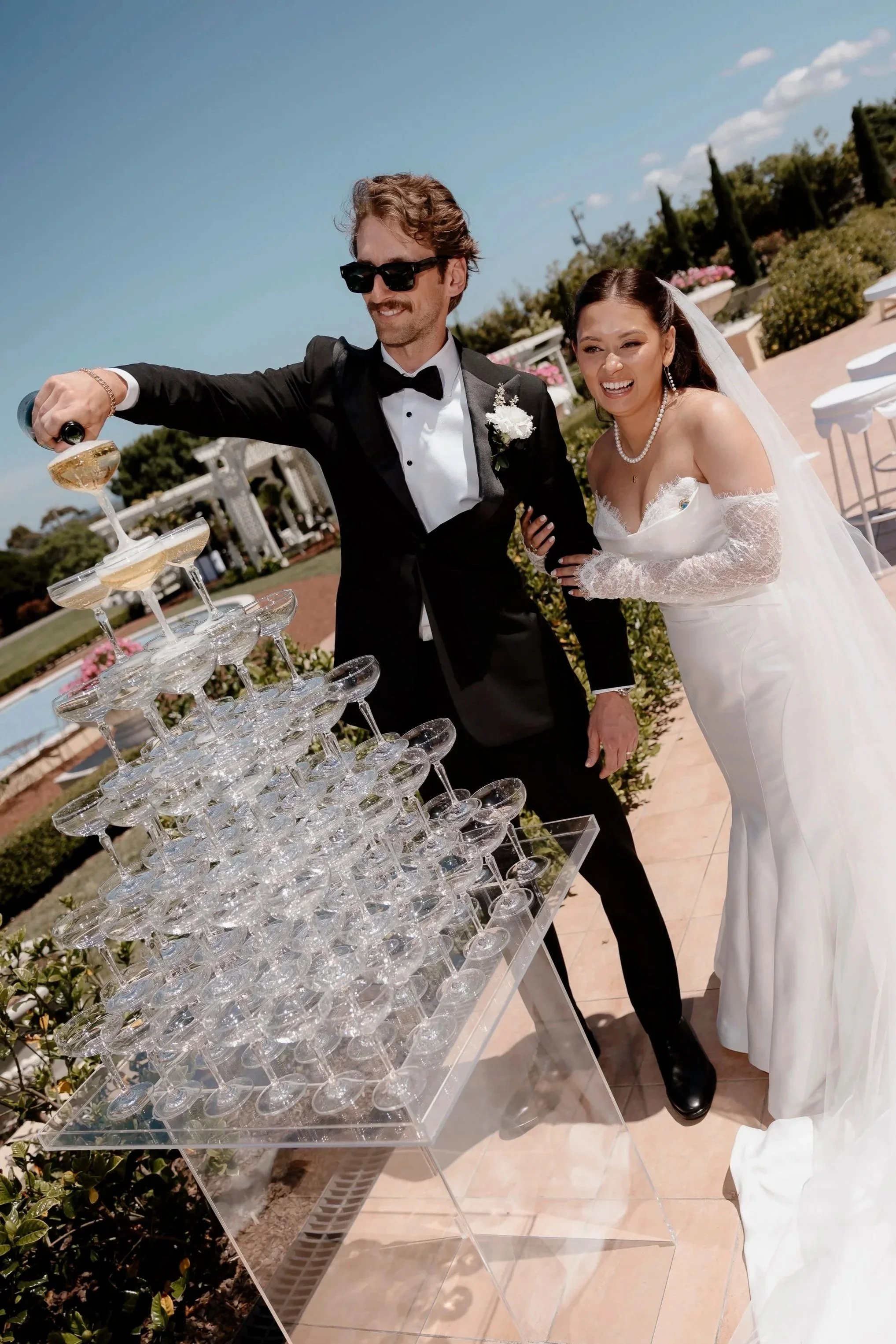 A bride and groom in wedding attire pour champagne into a pyramid of glasses during their wedding cocktail hour..