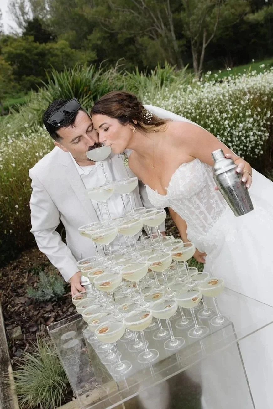 Bride and groom toasting with champagne over a pyramid of glasses at an outdoor wedding reception. Margarita Tower with shaker at cocktail hour