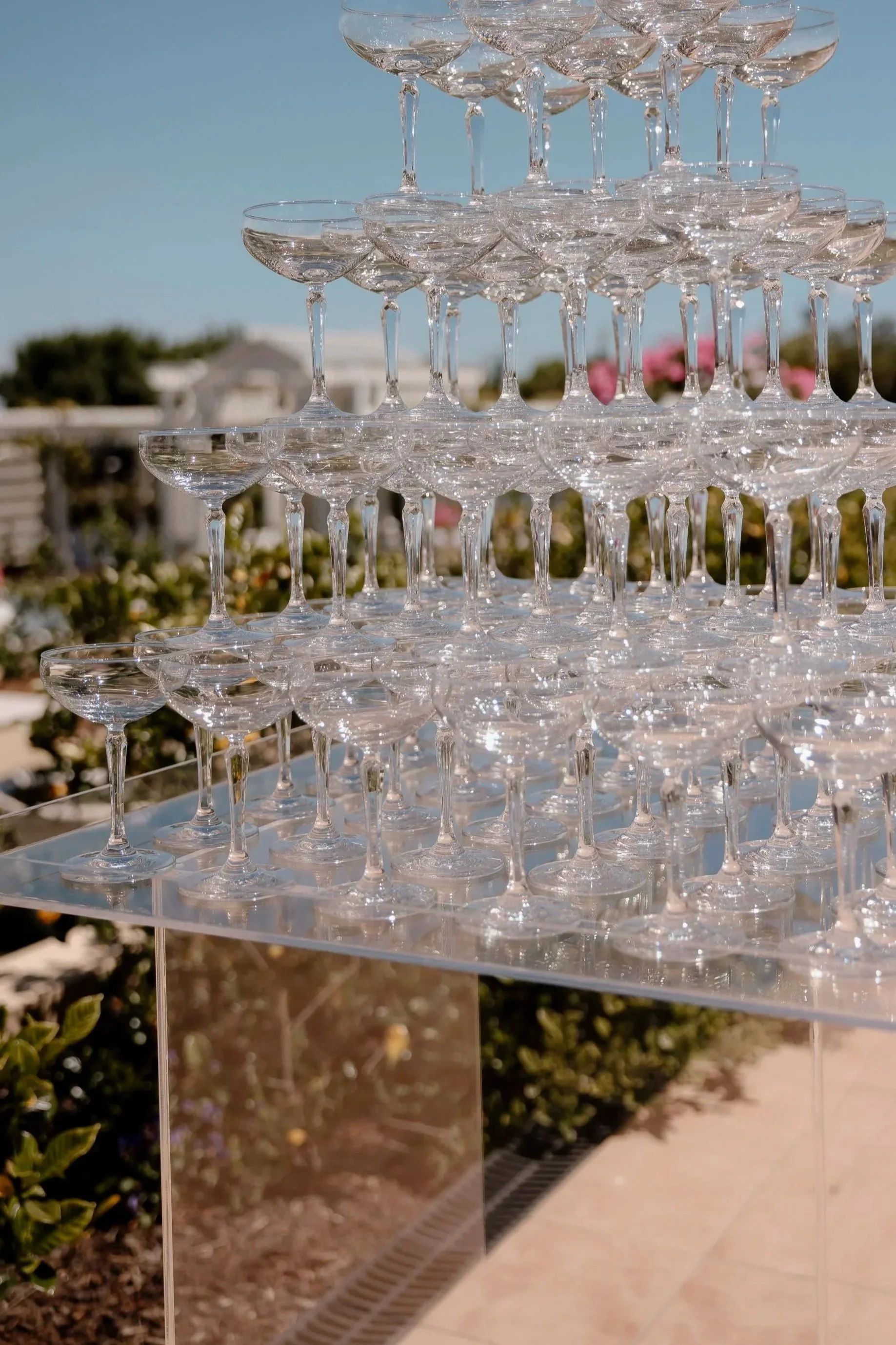 A large pyramid of empty champagne glasses arranged outdoors on a clear acrylic stand, with a blue sky and some pink flowering plants in the background.