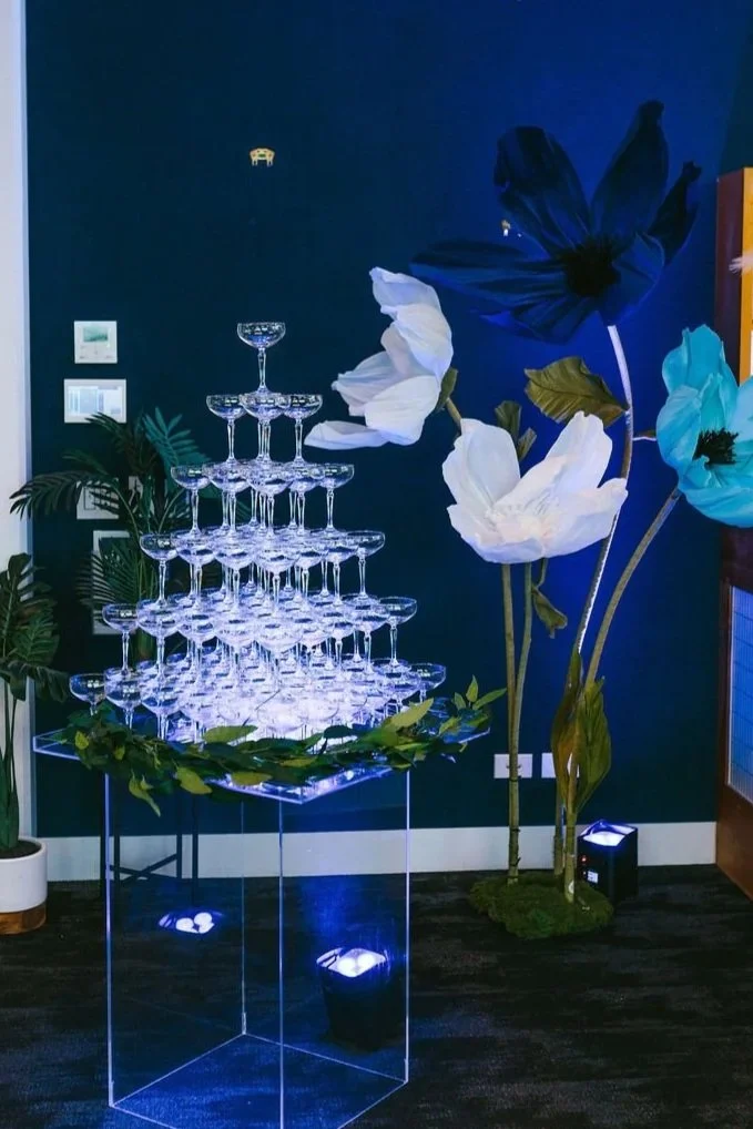A display of multiple champagne glasses arranged in a pyramid with white ribbons and bunches of green grapes, set on a clear stand at a wedding or celebration event.