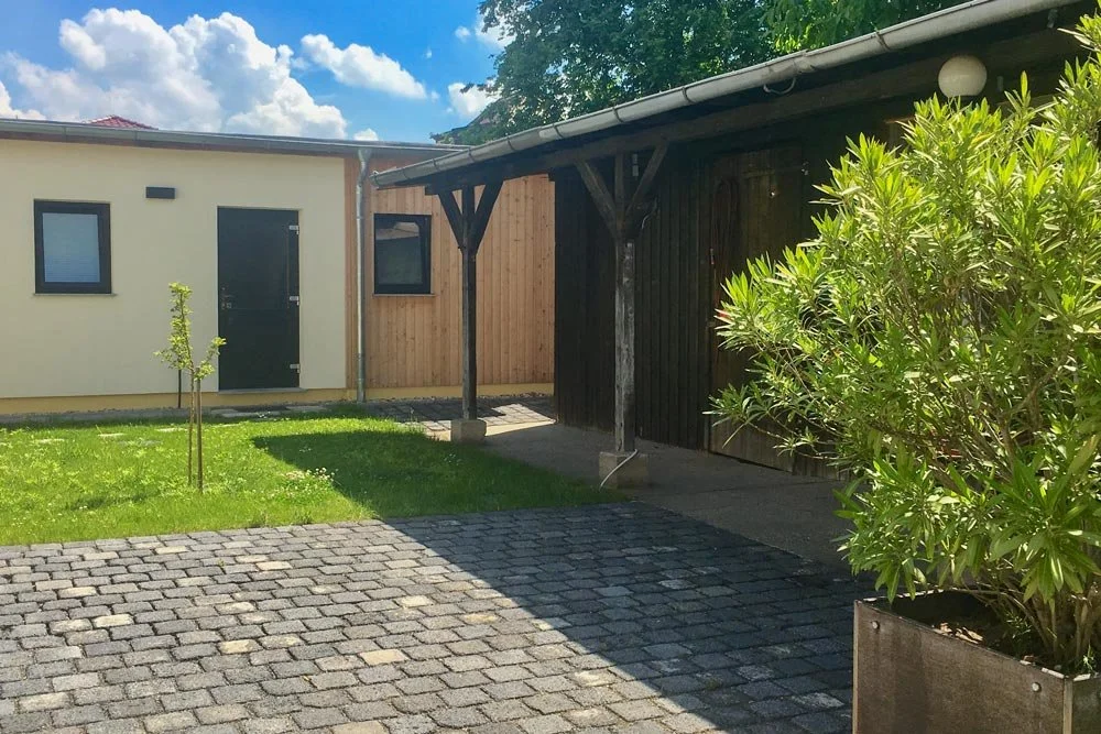  Blick auf den gepflasterten Hof und den grünen Außenbereich der Senfbude mit hellen Wänden und Holzelementen, ideal für Pausen von Tagesseminaren oder als Raum für Begegnung. 