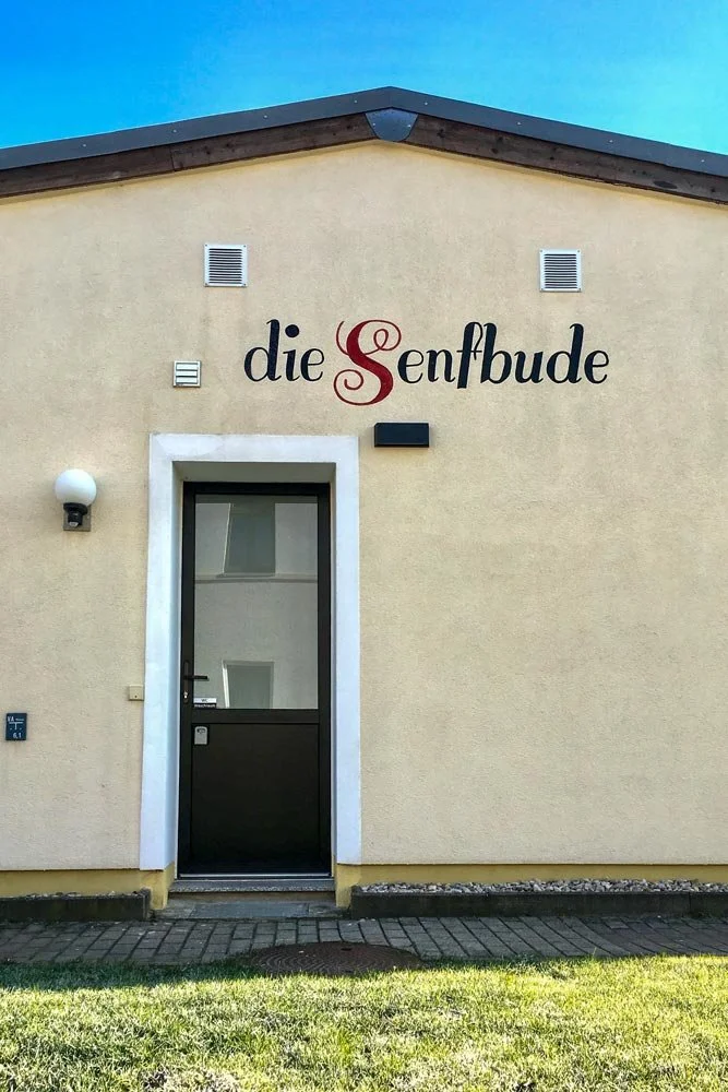  Außenansicht des Eingangs zum Seminar- &amp; Tagungsraum "die Senfbude" mit hellem Putz und dunkler Tür unter blauem Himmel. 