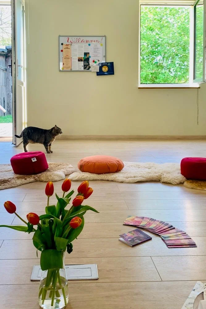  Ein einladender, heller Gruppenraum mit einer Tafel, Meditationskissen, warmen Fellen und einem Blumenstrauß. Eine Katze ist im Hintergrund zu sehen. Ein flexibler Raum für Austausch und Selbsterfahrung. 
