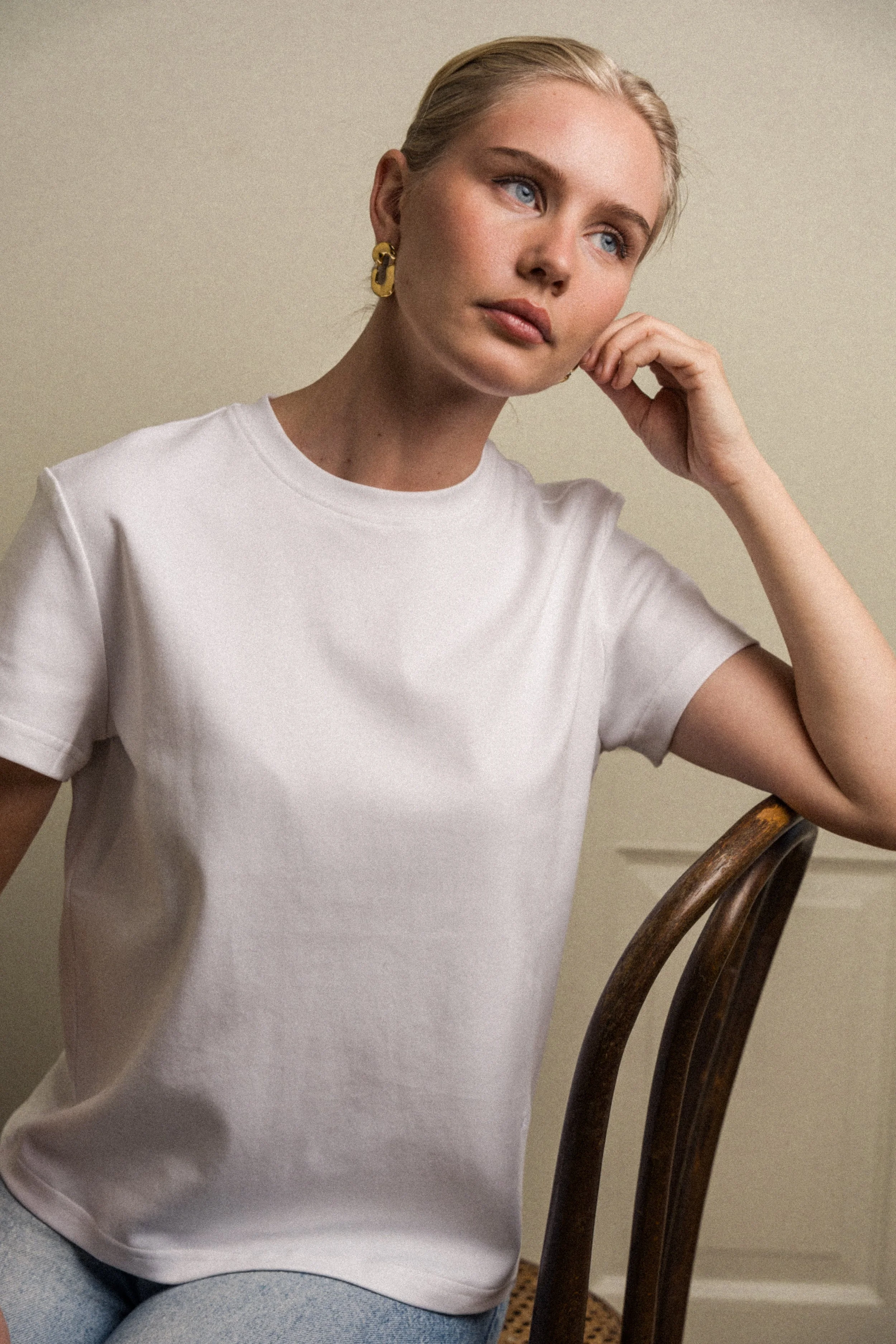 En kvinne med blondt hår og blå øyne sitter i en hvit t-skjorte, ser tenksom ut og hviler hodet på hånden, ved et trebord med en lysegrå bakgrunn.