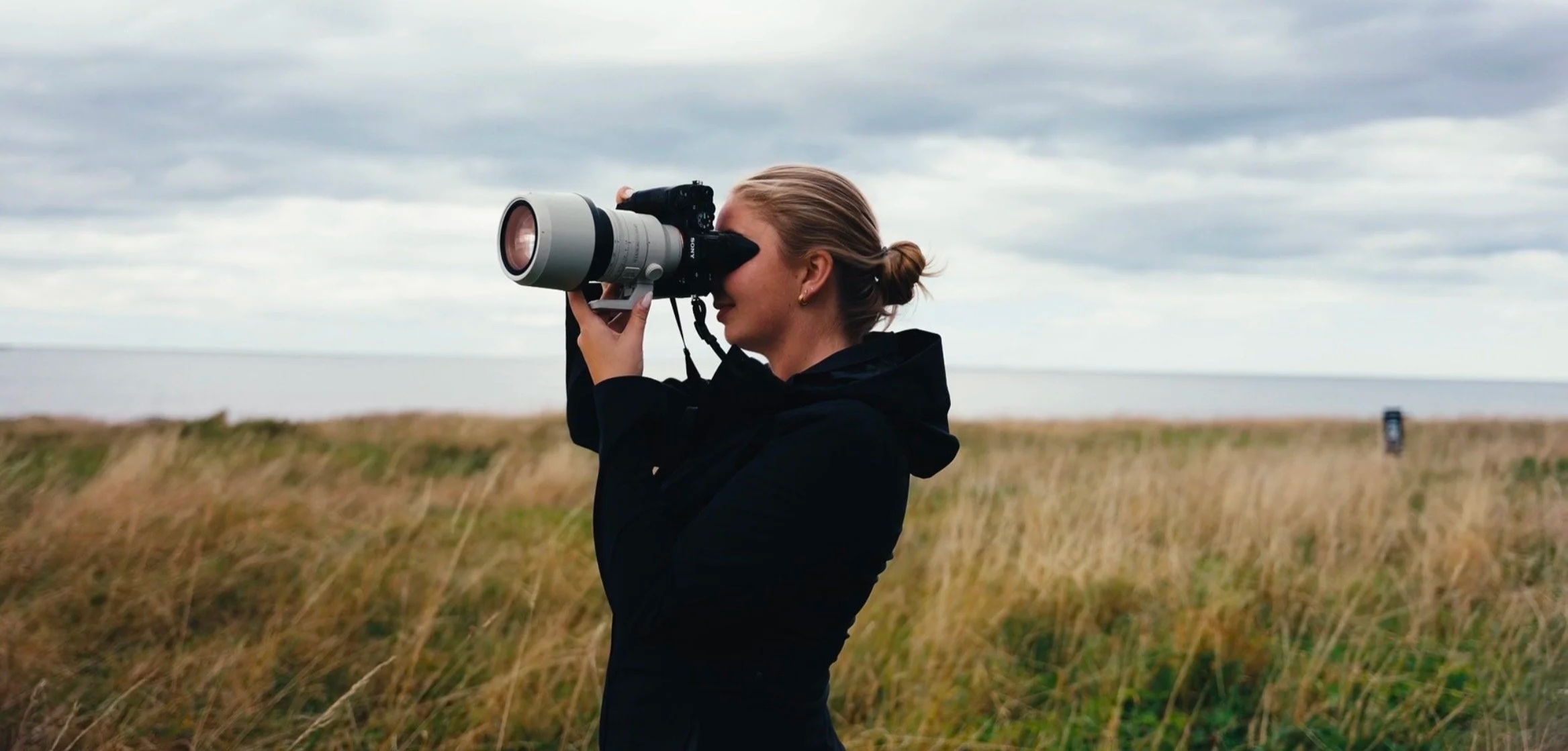 En kvinne ser gjennom et teleobjektiv med landskap av gressletter og hav i bakgrunnen.