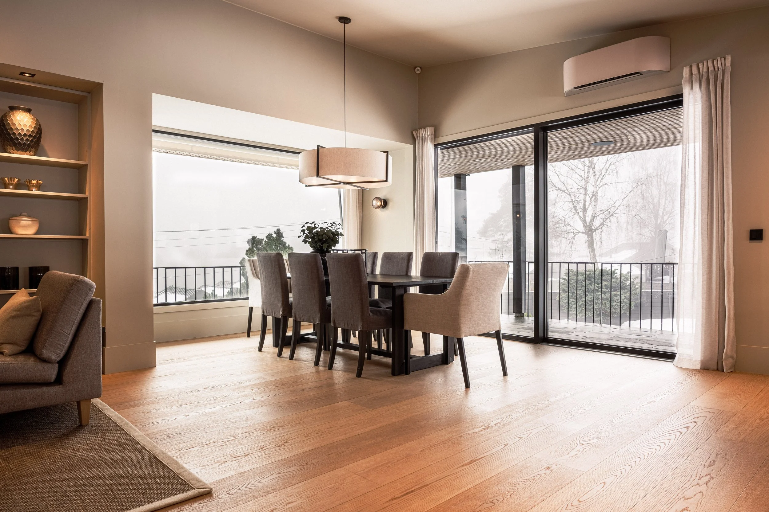 Stue med spisebord og åpen balkong med glassdører, store vinduer og tre utendørs trær.