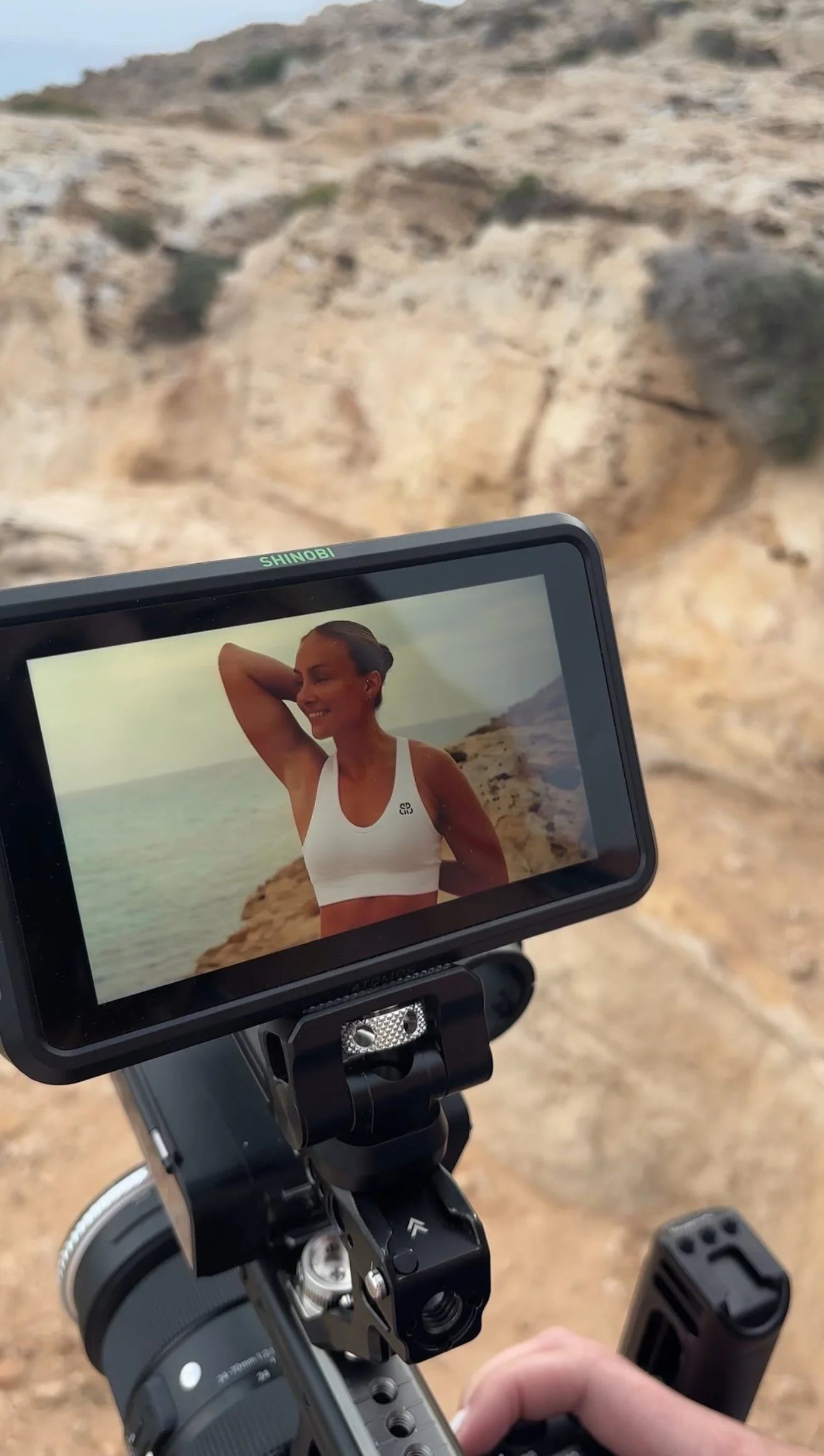 Et kameraskjermbilde av en kvinne på stranden, smilende, med hånden bak hodet, iført en hvit treningsbikini, med kystlinje i bakgrunnen.