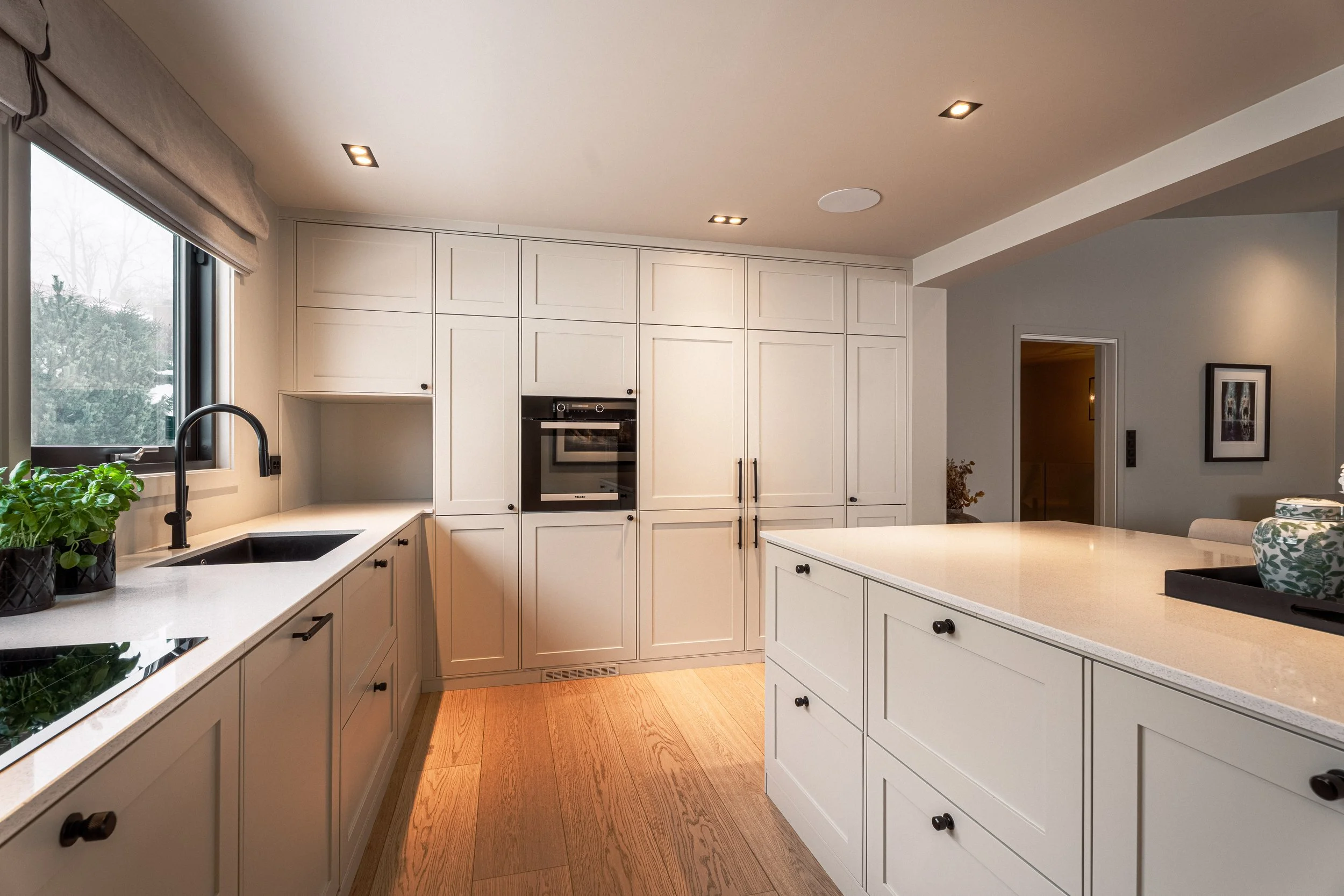 Moderne kjøkken med hvite skap, gryte under vindu, stekepanne på benkeplate, og kjøkkenøy i midten.