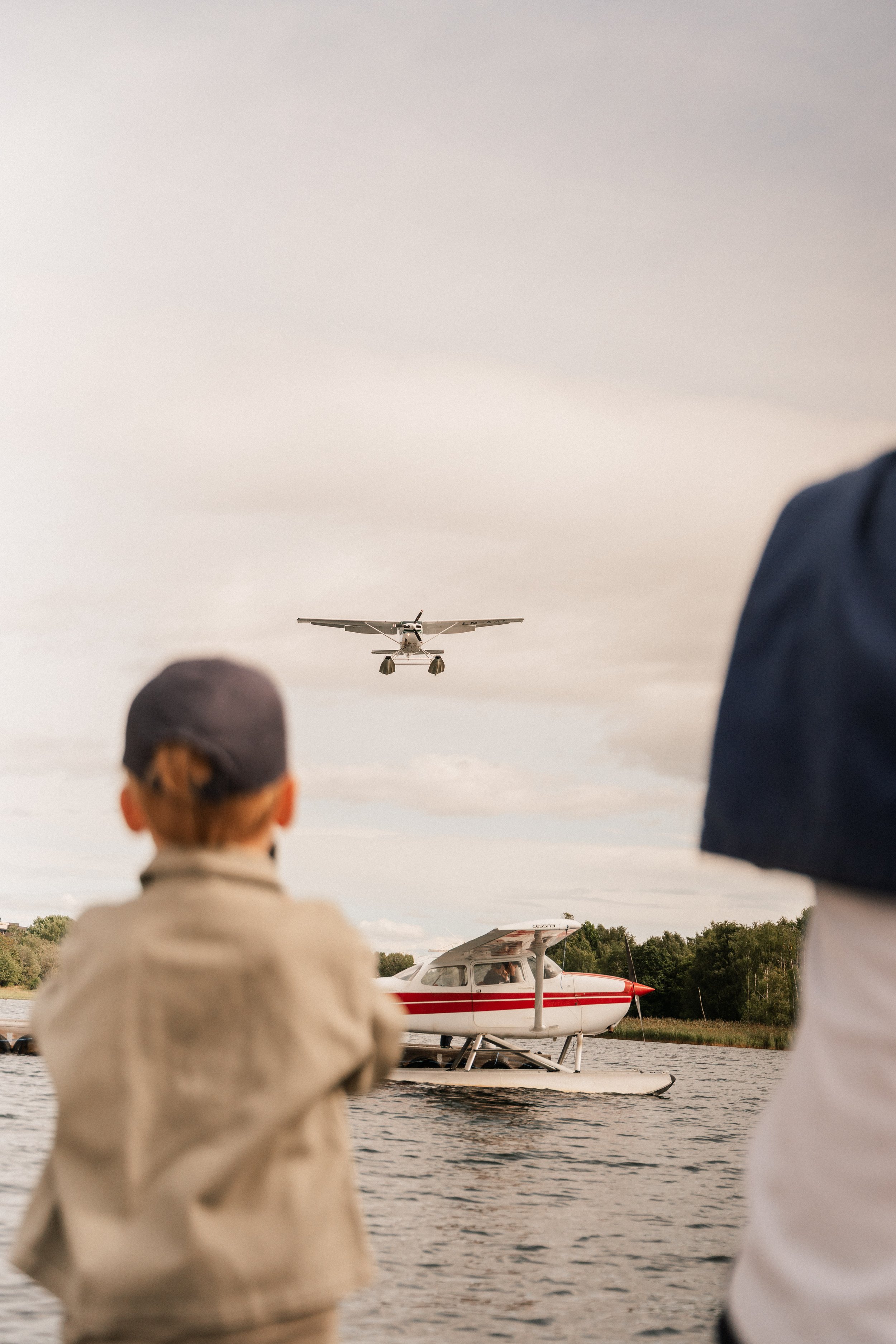 Personer ser på en flygende drone over vannet, med båter og fly på bakken i bakgrunnen.