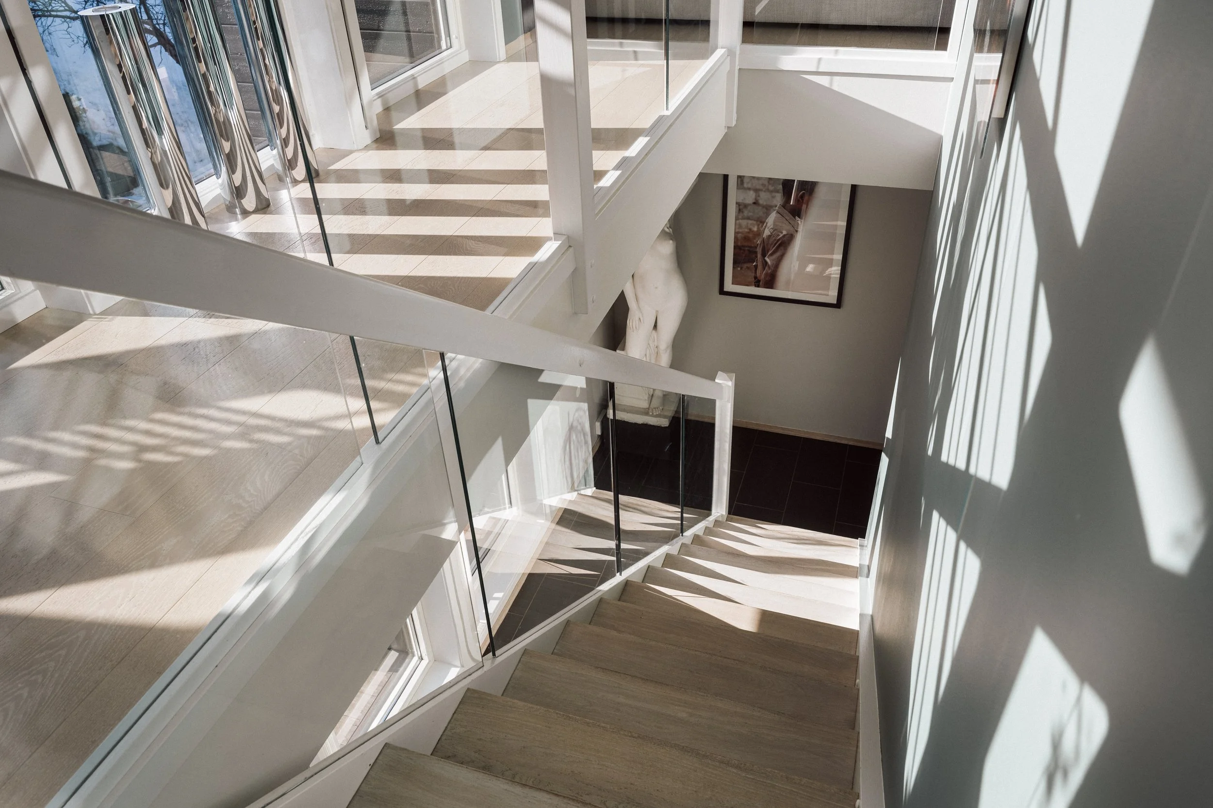 Innendørs trapp med trappetrinn av tre og glassrekkverk, sollys som kaster skygger gjennom vinduer, og en vegg med et bilde og en mannlig statue.