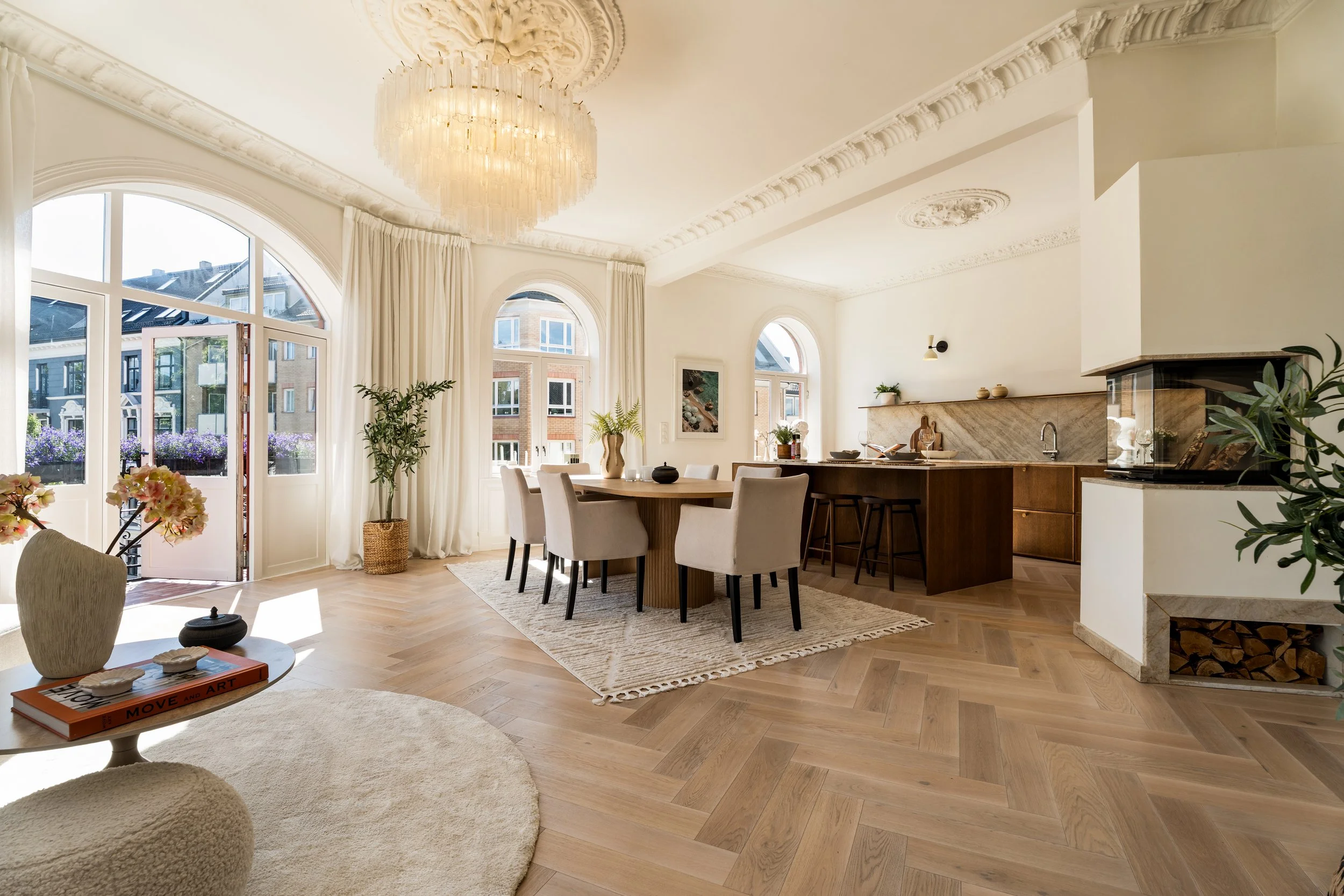 Stue med beige sofa, armlene, puter, lenestol, sofa, stuebord, TV, vindu med gardiner, trestamme, kunstverk, peis, spisebord, kjøkken i bakgrunnen.