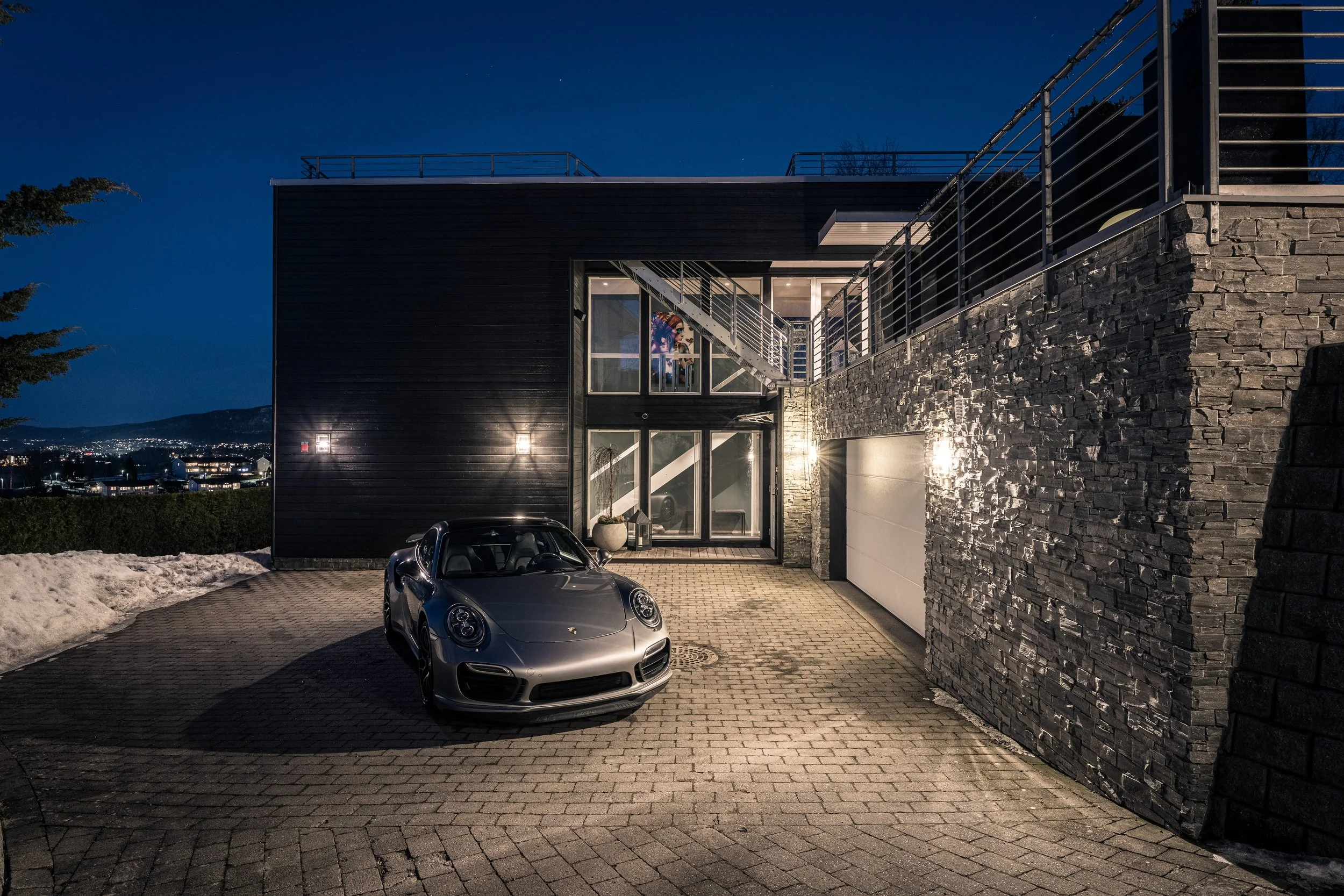 Moderne svart hus med en parkeringsplass foran, en sølvbil parkert og utendørsbelysning som lyser opp veggen, nattestid.