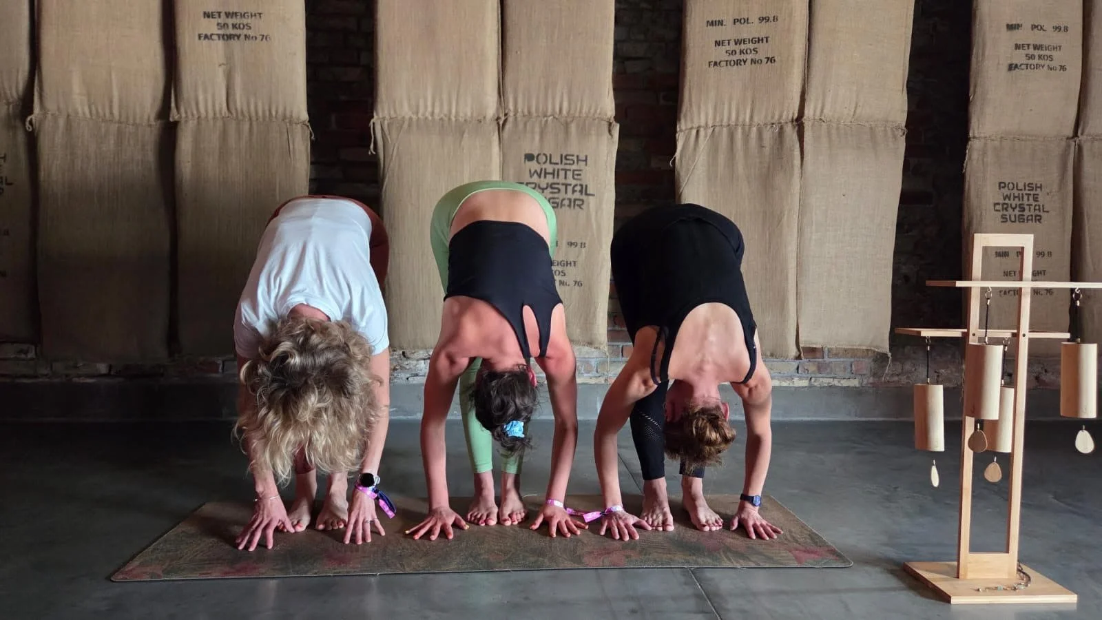 Four women practicing yoga in a downward dog pose on a mat, in a room with large sacks in the background and a wooden percussion instrument to the right.
