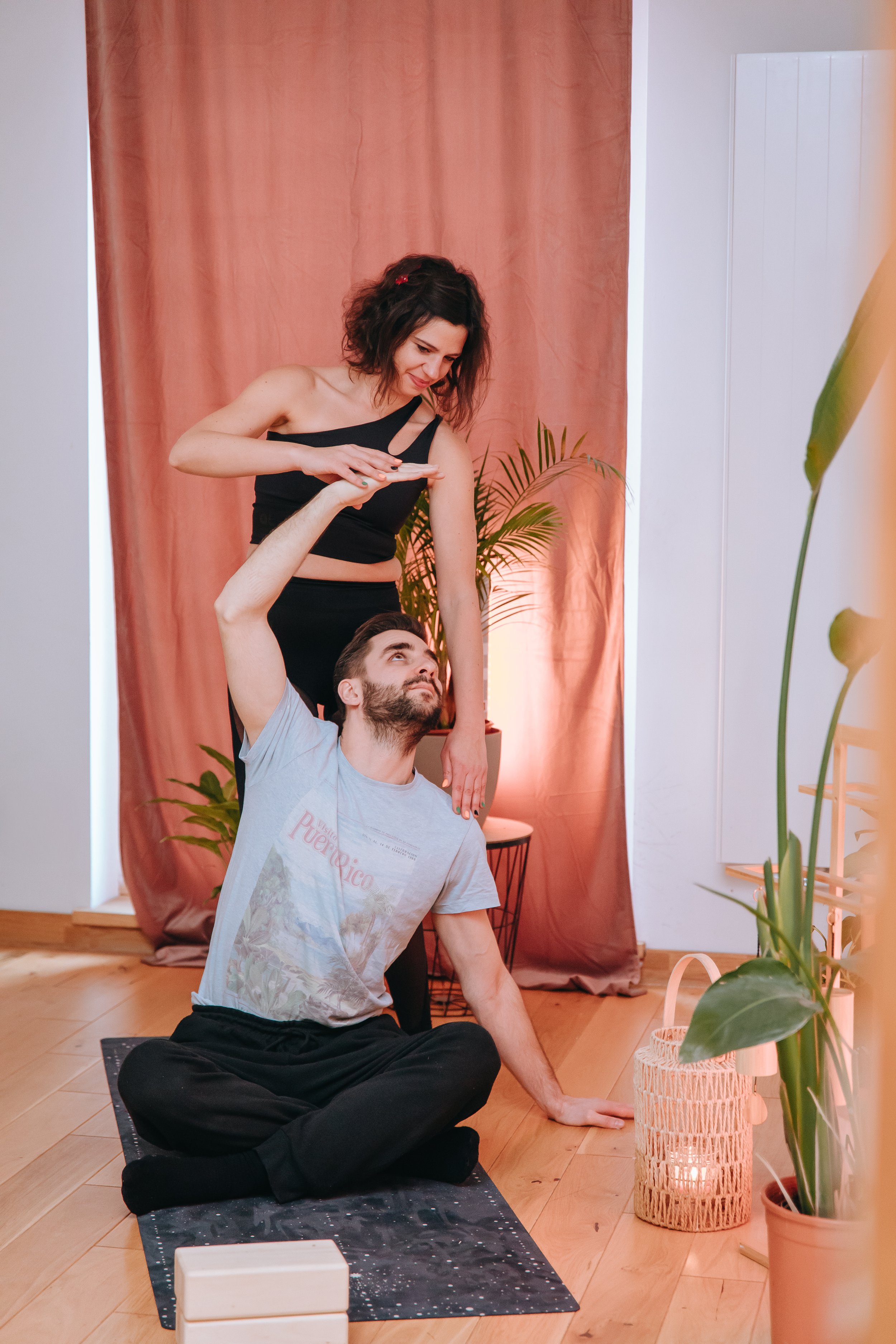 A woman in a black top and a man in a gray t-shirt are practicing acro yoga in a cozy room with wooden floors, pink curtains, and potted plants. The woman stands on her hands on the man's knees as he sits cross-legged on the floor, looking up at her.