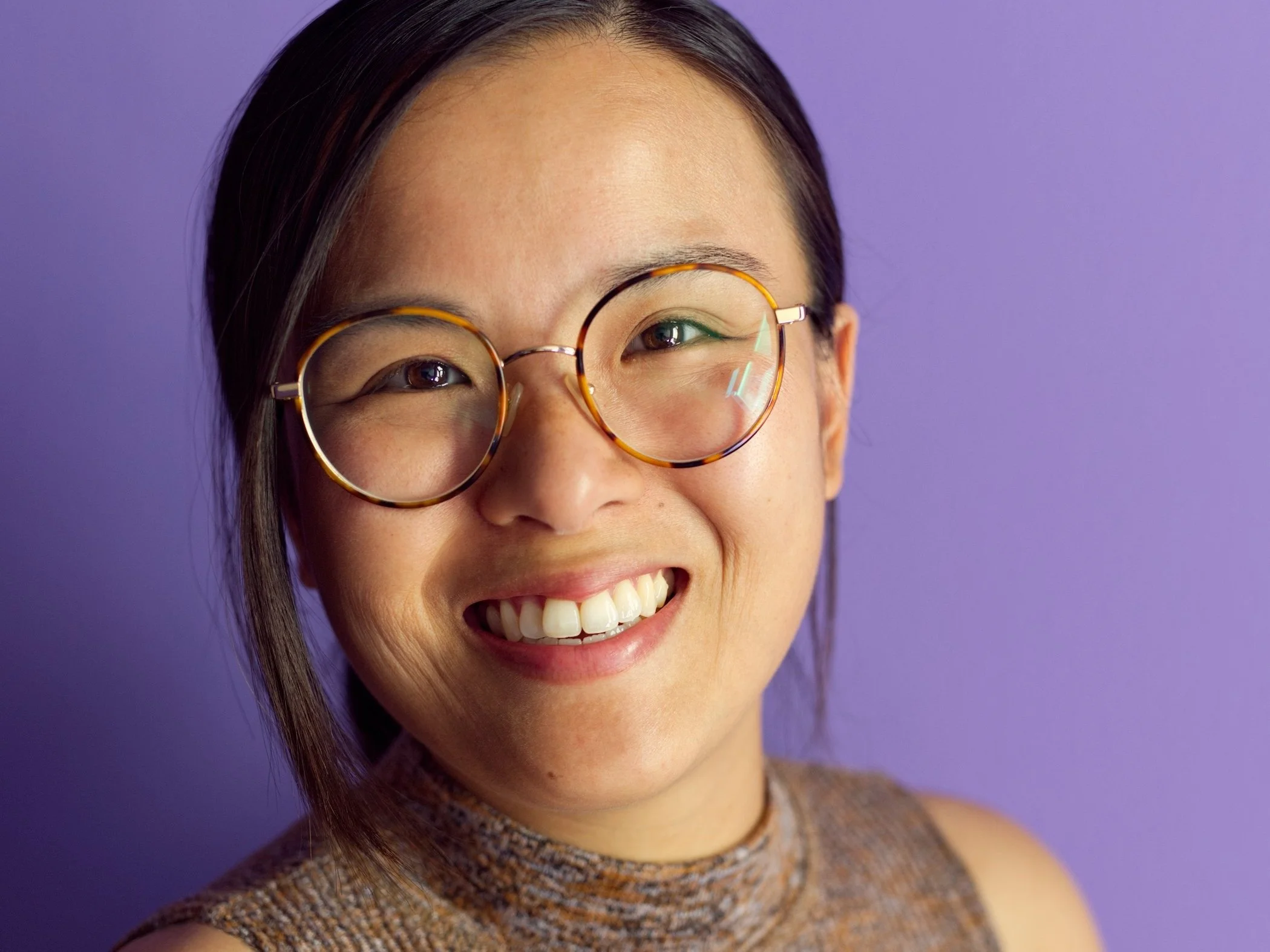 A woman smiling, wearing round tortoiseshell glasses and a sleeveless, high-neck, patterned top, standing against a purple background.