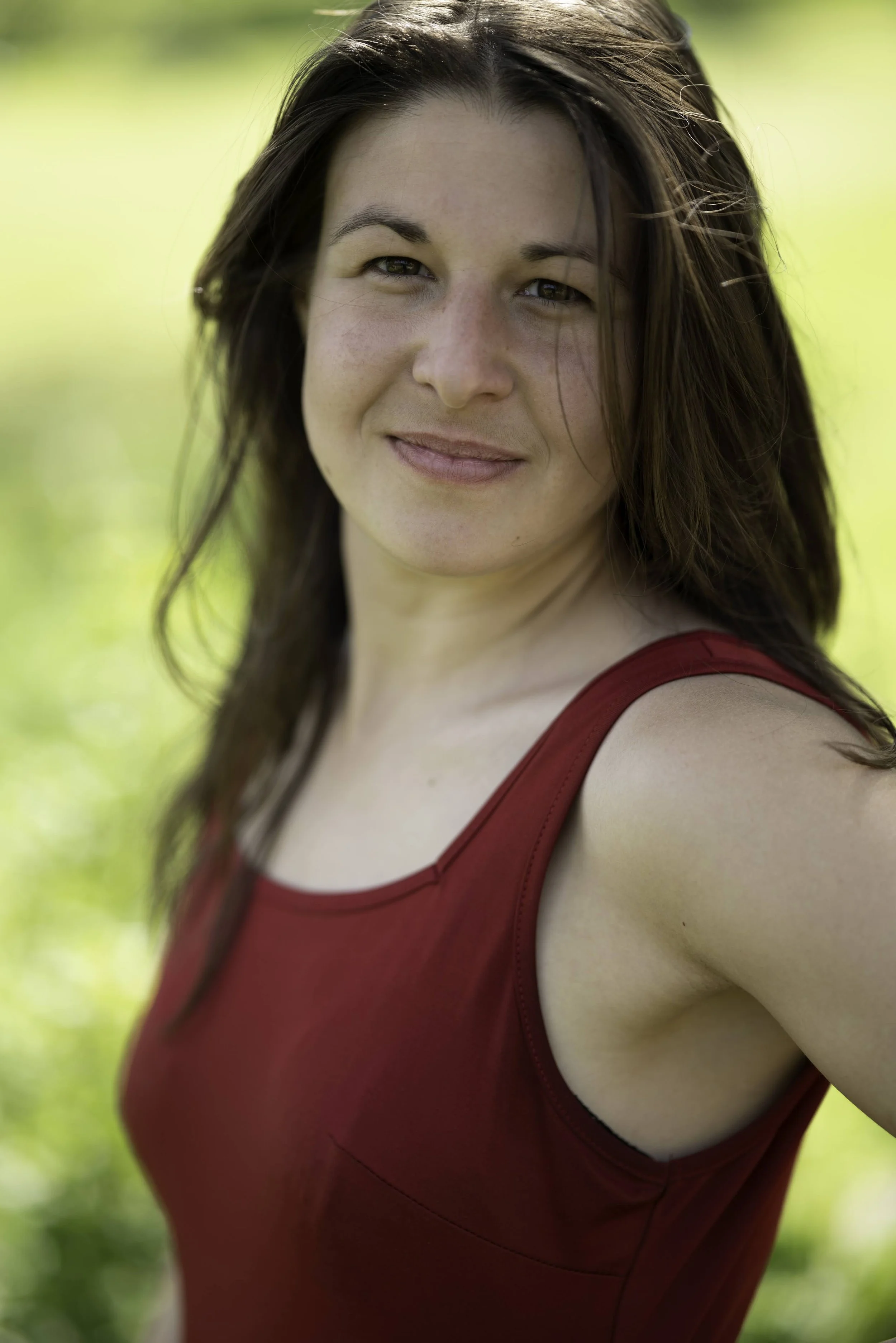 A woman with brown hair wearing a red sleeveless top outdoors with a grassy background.