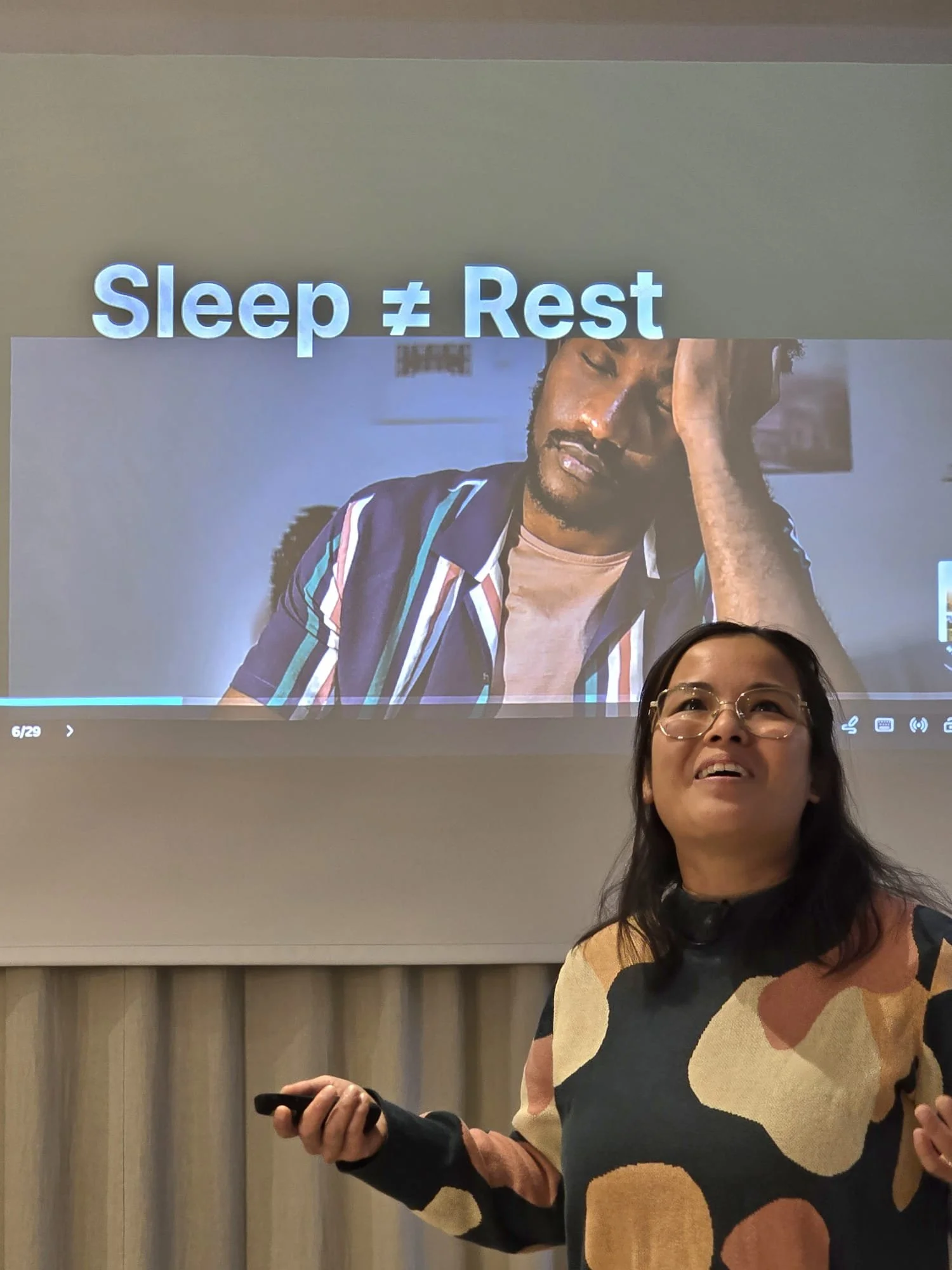 A woman giving a presentation in a room with a large screen behind her. The screen displays the text "Sleep ≠ Rest" and a photo of a man with a tired expression, resting his head on his hand.